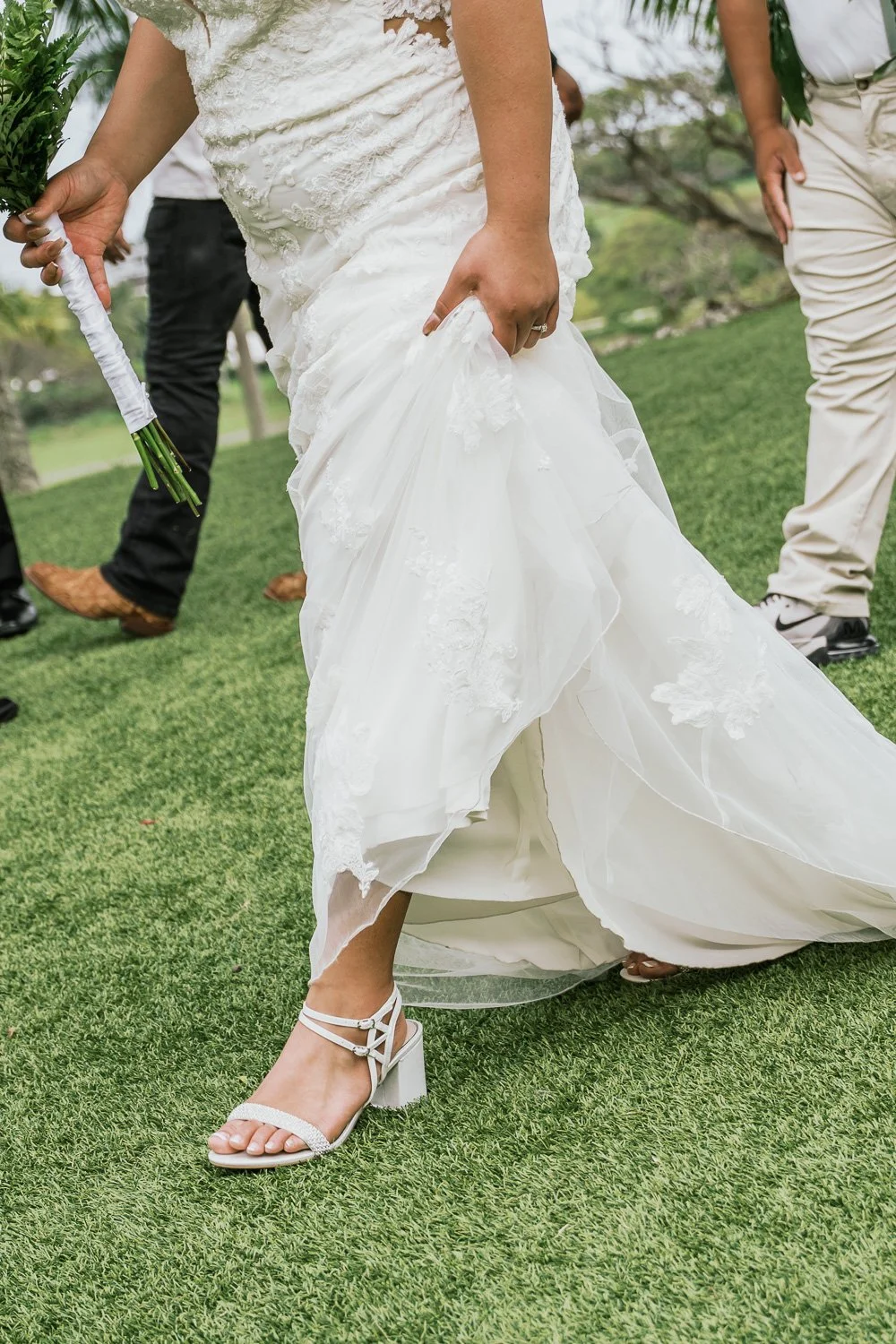 A bride in a white lace wedding gown and white high-heeled shoes lifts her dress while walking on a grassy outdoor area, with guests in the background.