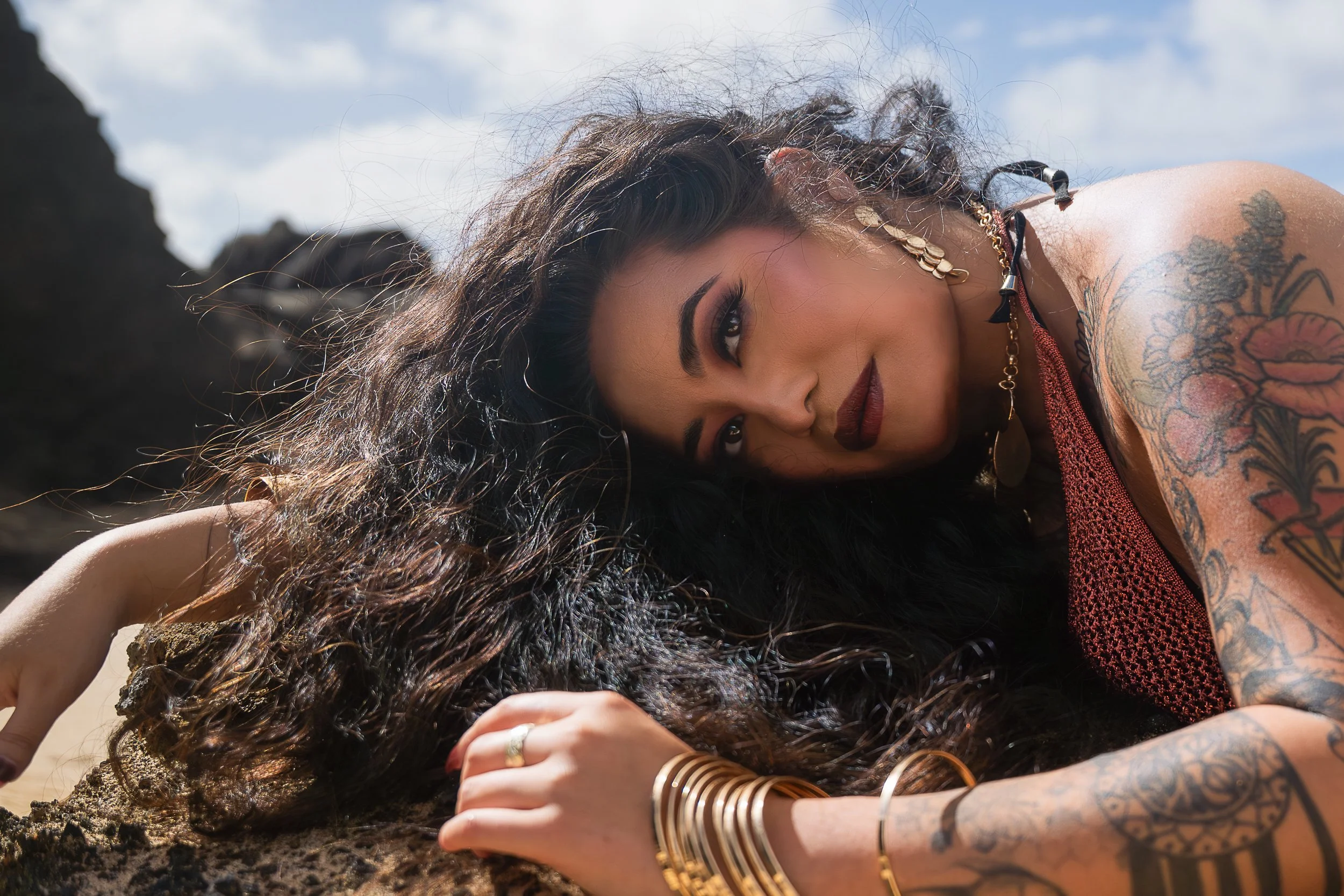 A woman with long curly black hair, tattoos on her arms, and gold jewelry lying on her side outdoors with a rocky background and cloudy sky.