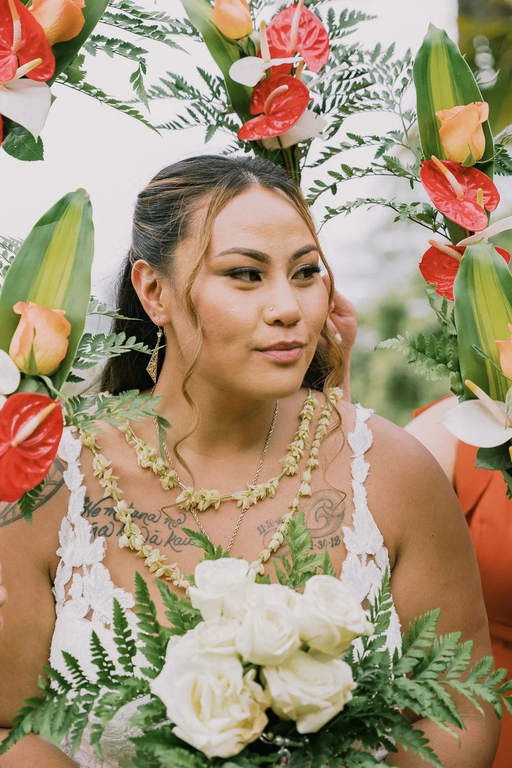 A woman with long brown hair, earrings, and tattoos, wearing a white lace dress, holds a bouquet of white roses and greenery, framed by tropical flowers, during a celebration or ceremony outdoors.