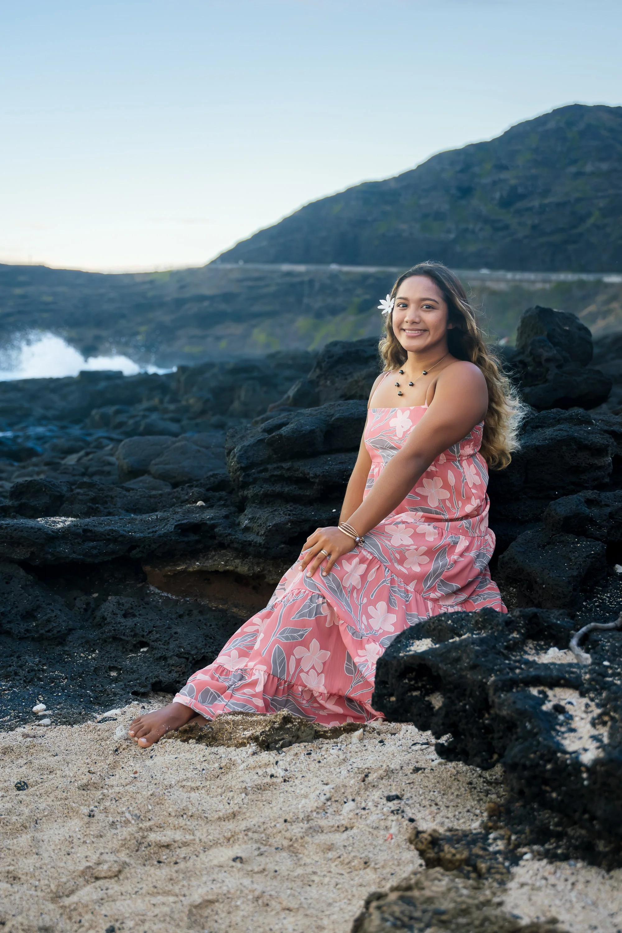 A young woman in a pink floral dress sitting on volcanic rocks at the beach, smiling at the camera with hills and ocean in the background.