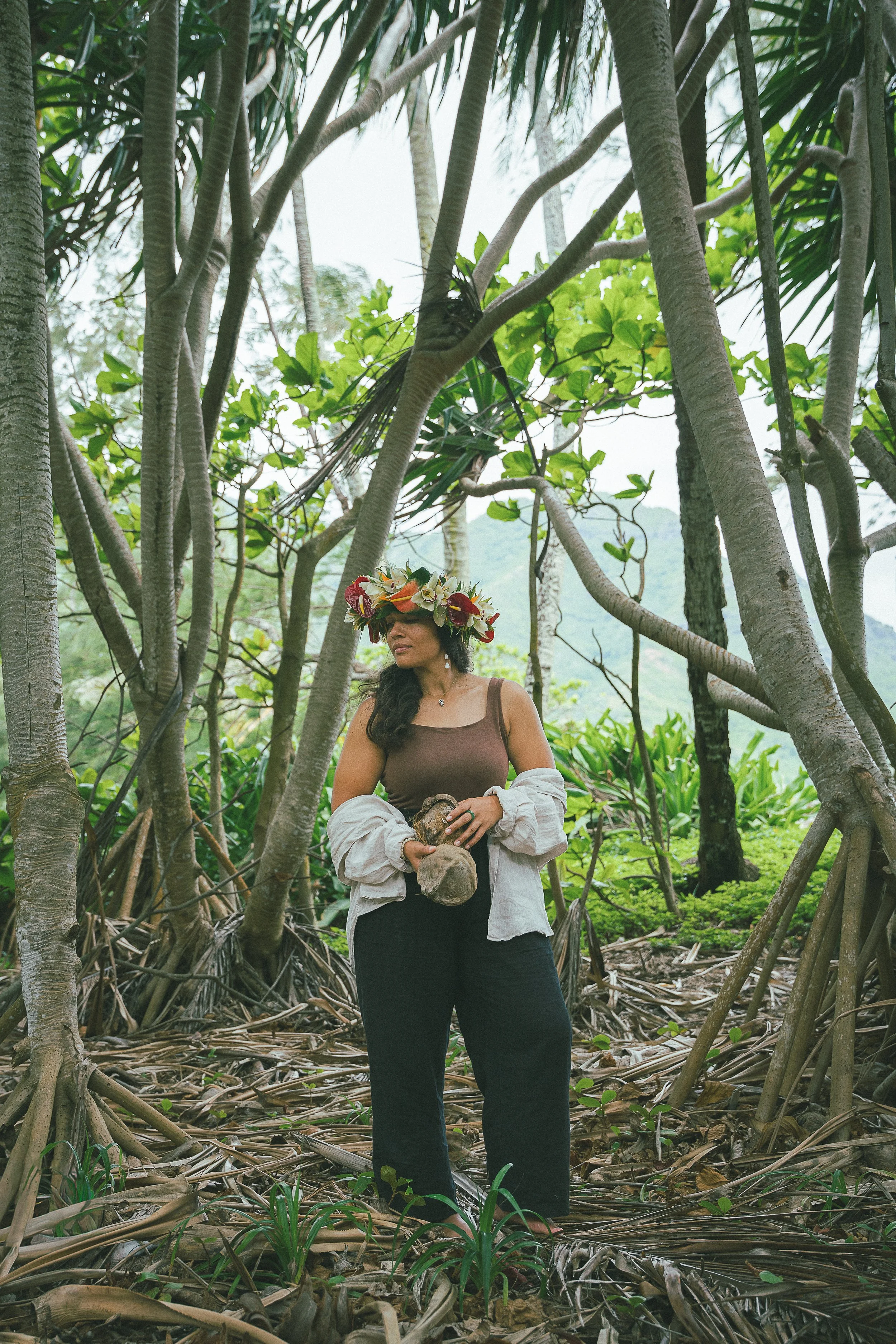 A woman wearing a floral crown, tank top, and a white jacket draped over her arms stands in a dense tropical forest holding a piece of wood.