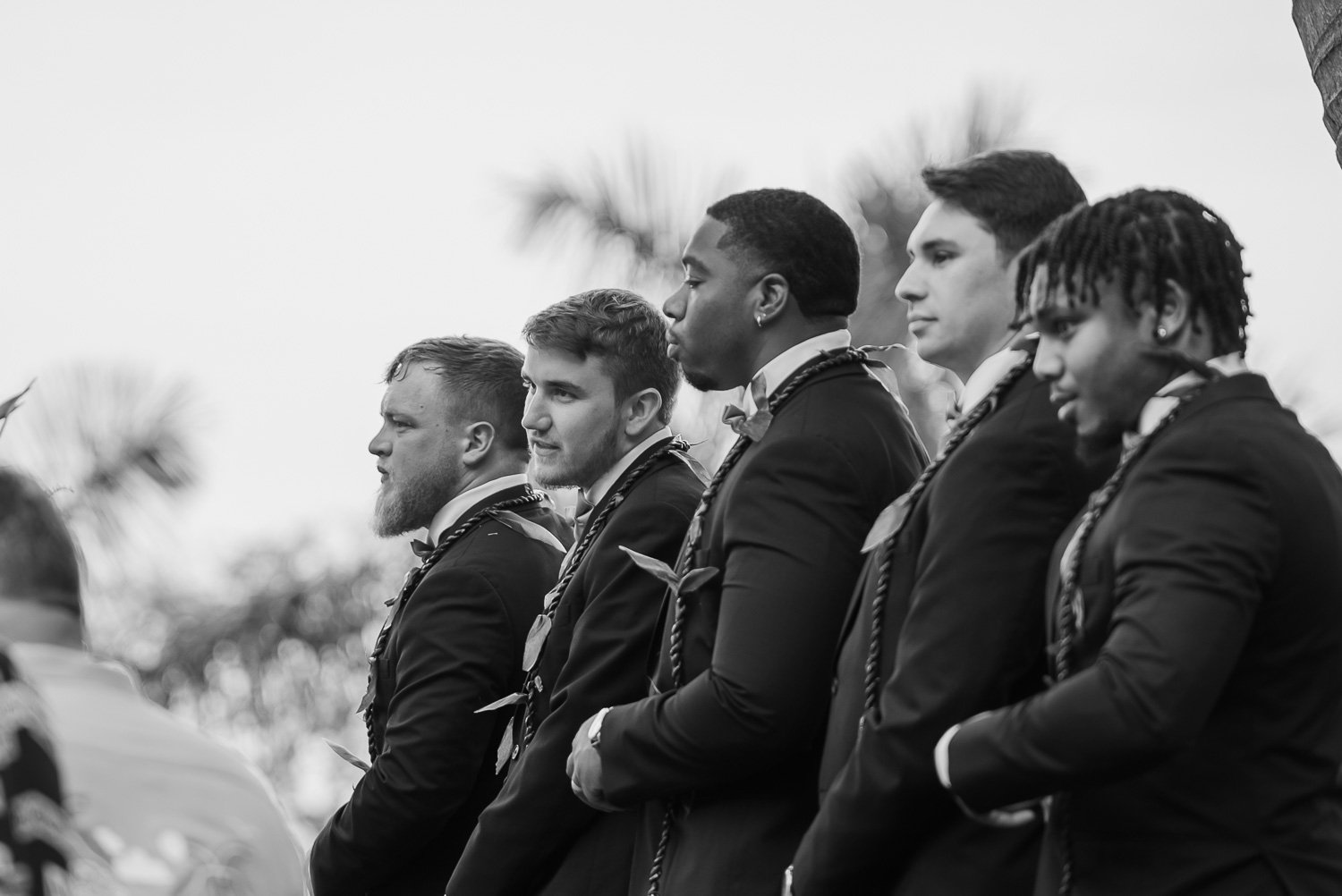 A group of young men dressed in formal suits and accessories, standing in a line outdoors, possibly at a formal event or ceremony, with palm trees in the background.