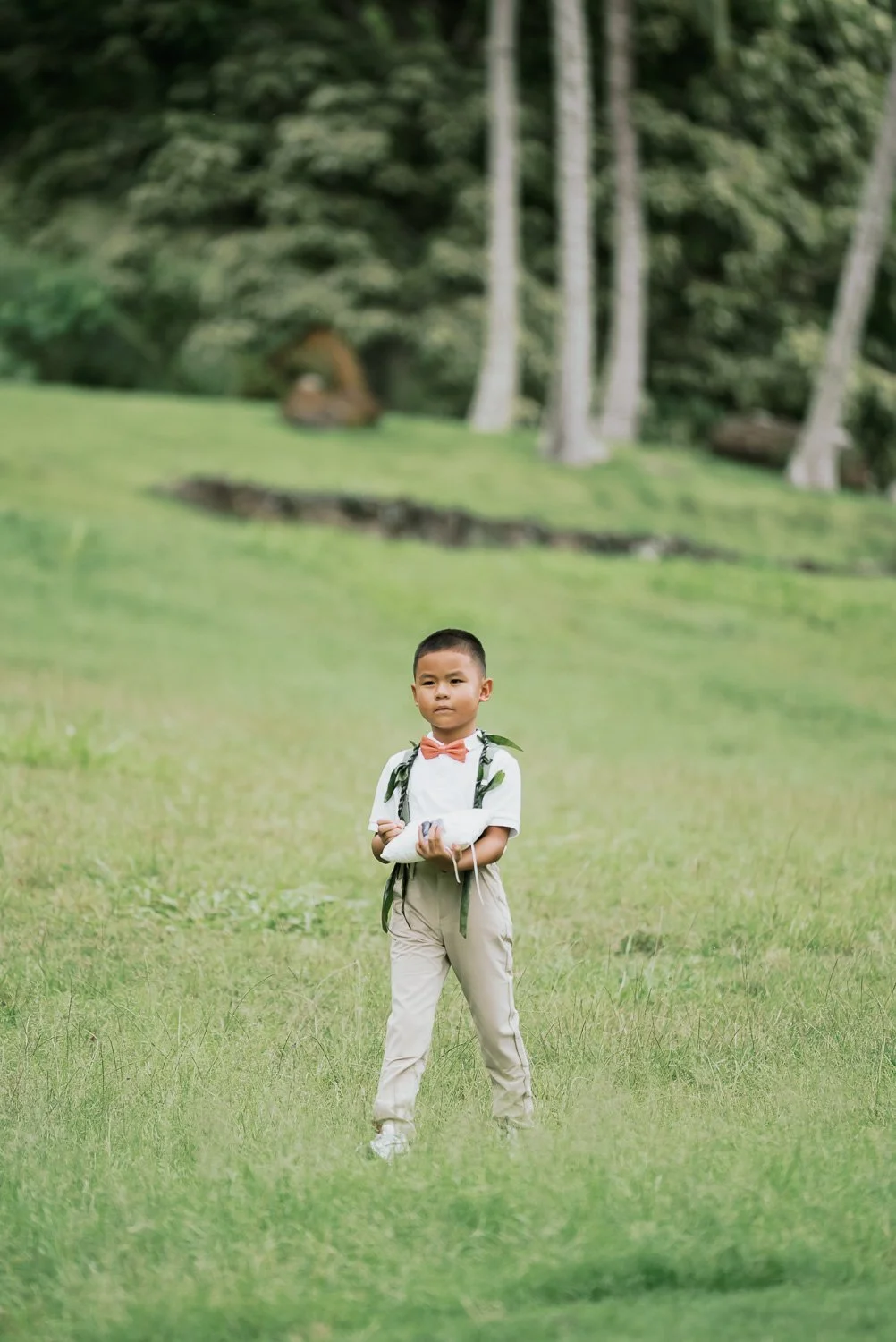 A young boy standing on a grassy field with trees in the background, wearing a white shirt, beige pants, and a pink bow tie, holding a white envelope.