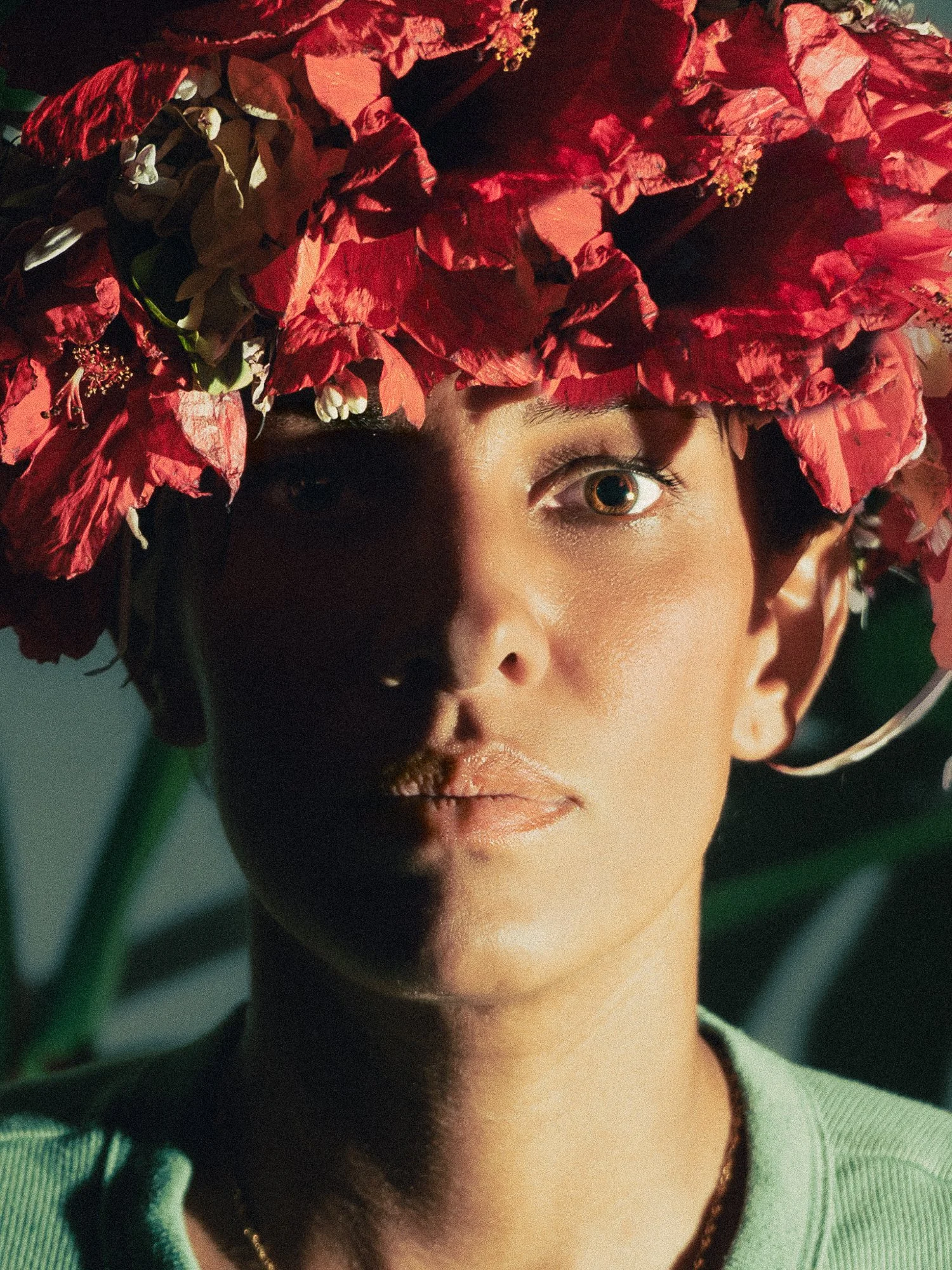 A woman with a large crown of red and pink flowers on her head, with one eye partially shadowed.