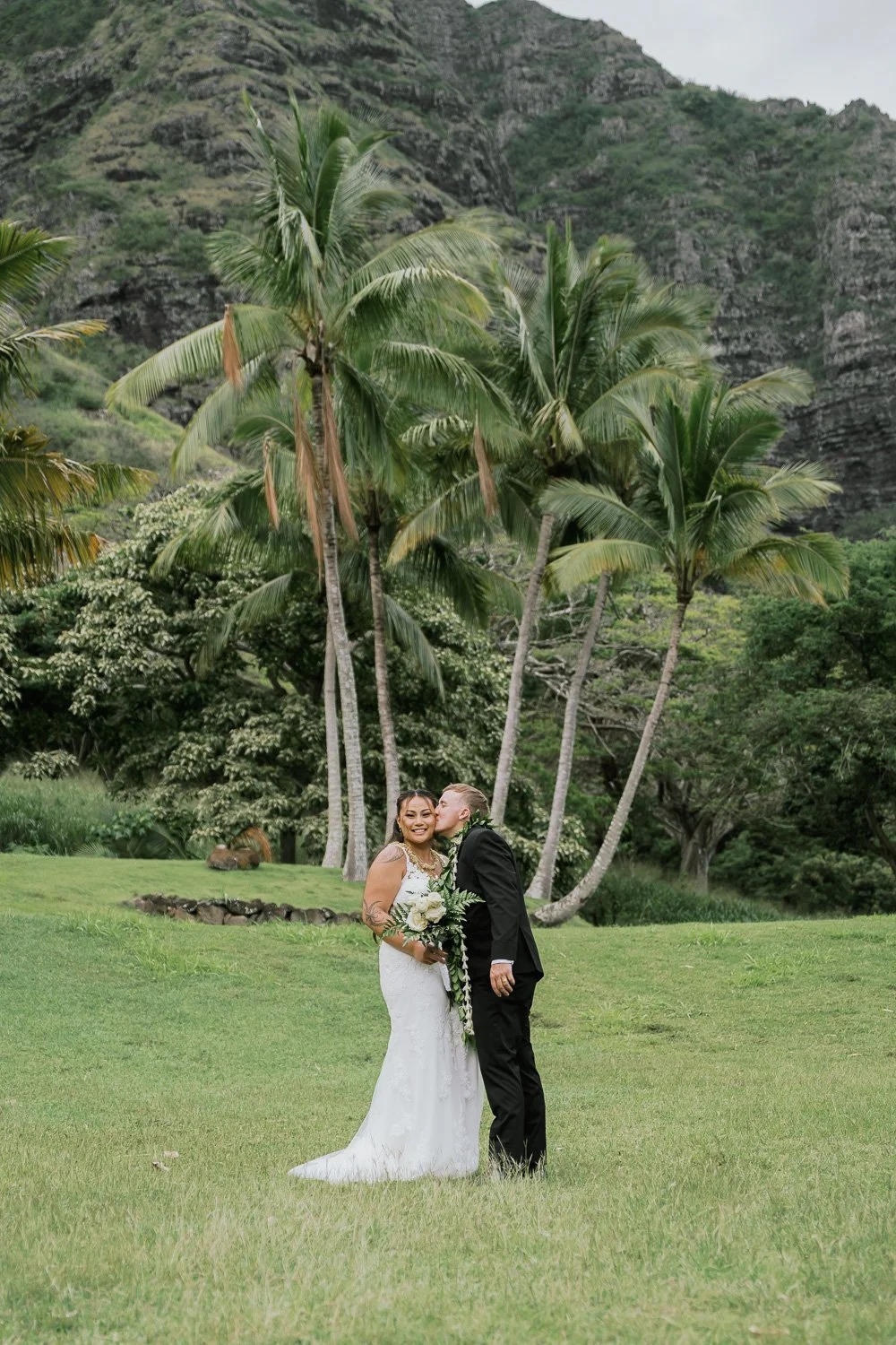A bride and groom sharing a kiss on a lush green lawn with tropical palm trees and mountainous terrain in the background.