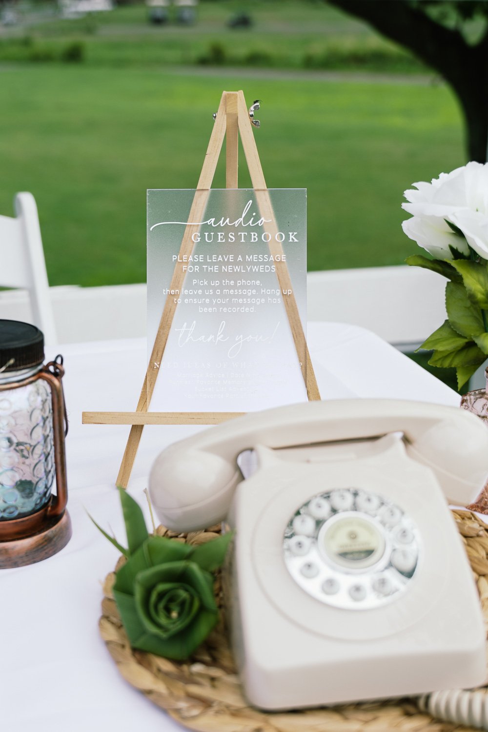 A vintage rotary telephone on a table with a small green succulent plant, part of a guestbook sign about leaving messages for newlyweds, and floral decorations, set outdoors in a garden or park.