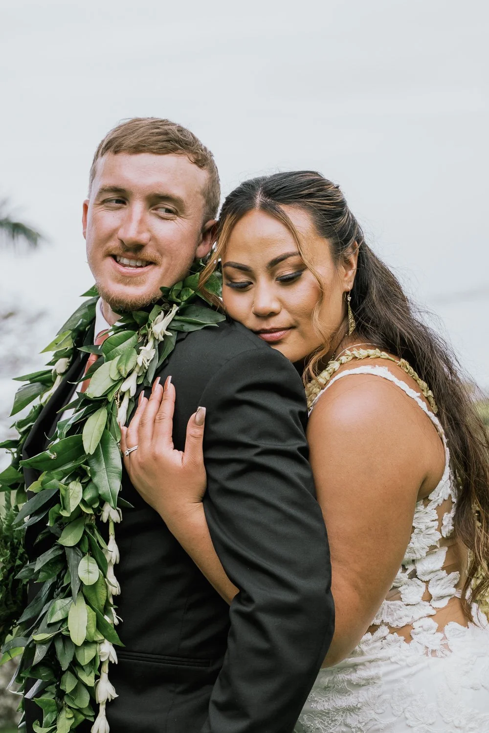 A bride and groom embracing outdoors on their wedding day. The groom is smiling and wearing a black suit with a leafy lei around his neck. The bride, with long dark hair, is wearing a white lace dress and looks peaceful as she leans on the groom's sh