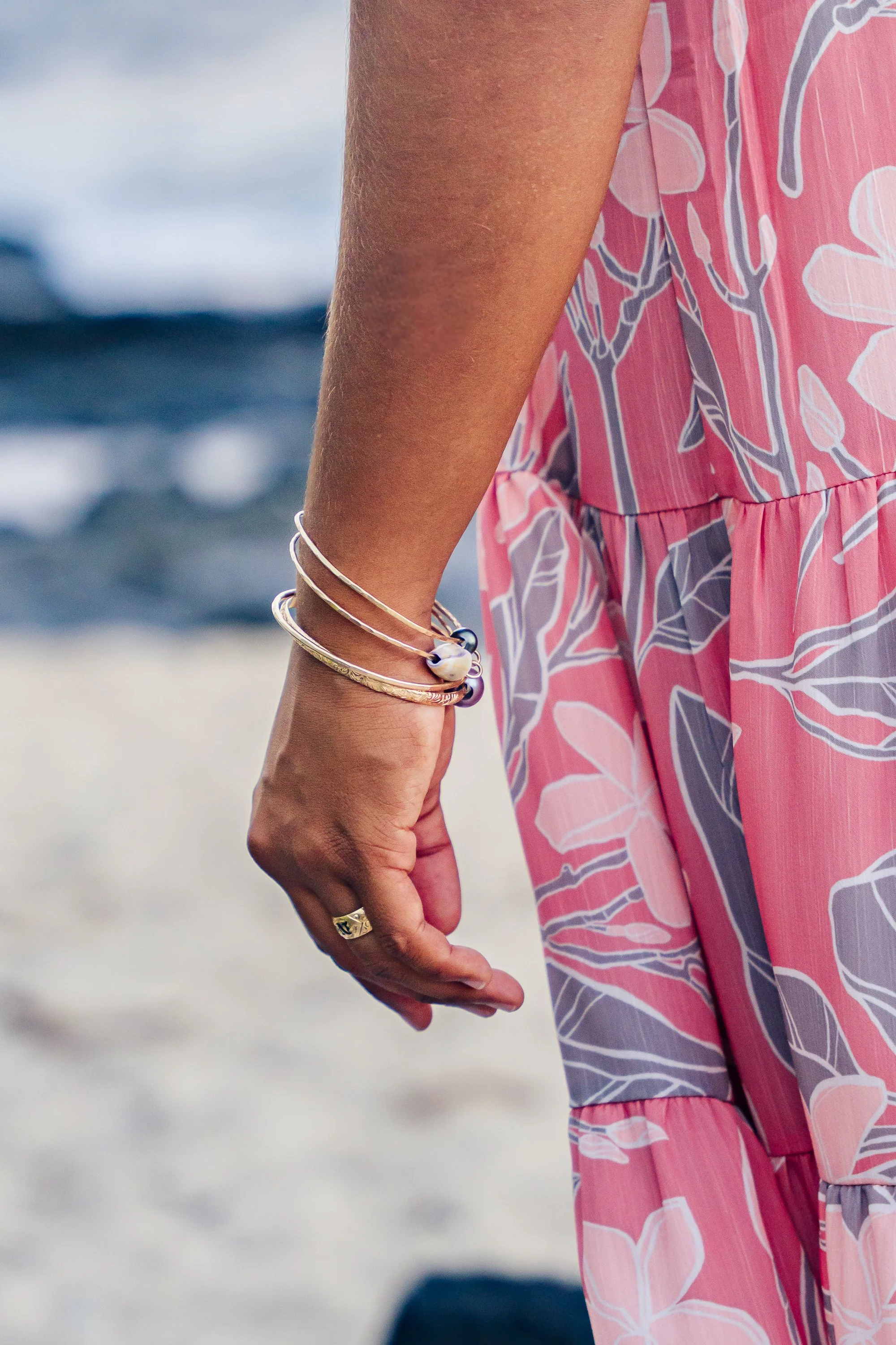 Close-up of a woman's arm and hand at the beach, wearing multiple silver bracelets and a ring, dressed in a pink and gray floral dress, with ocean waves in the background.