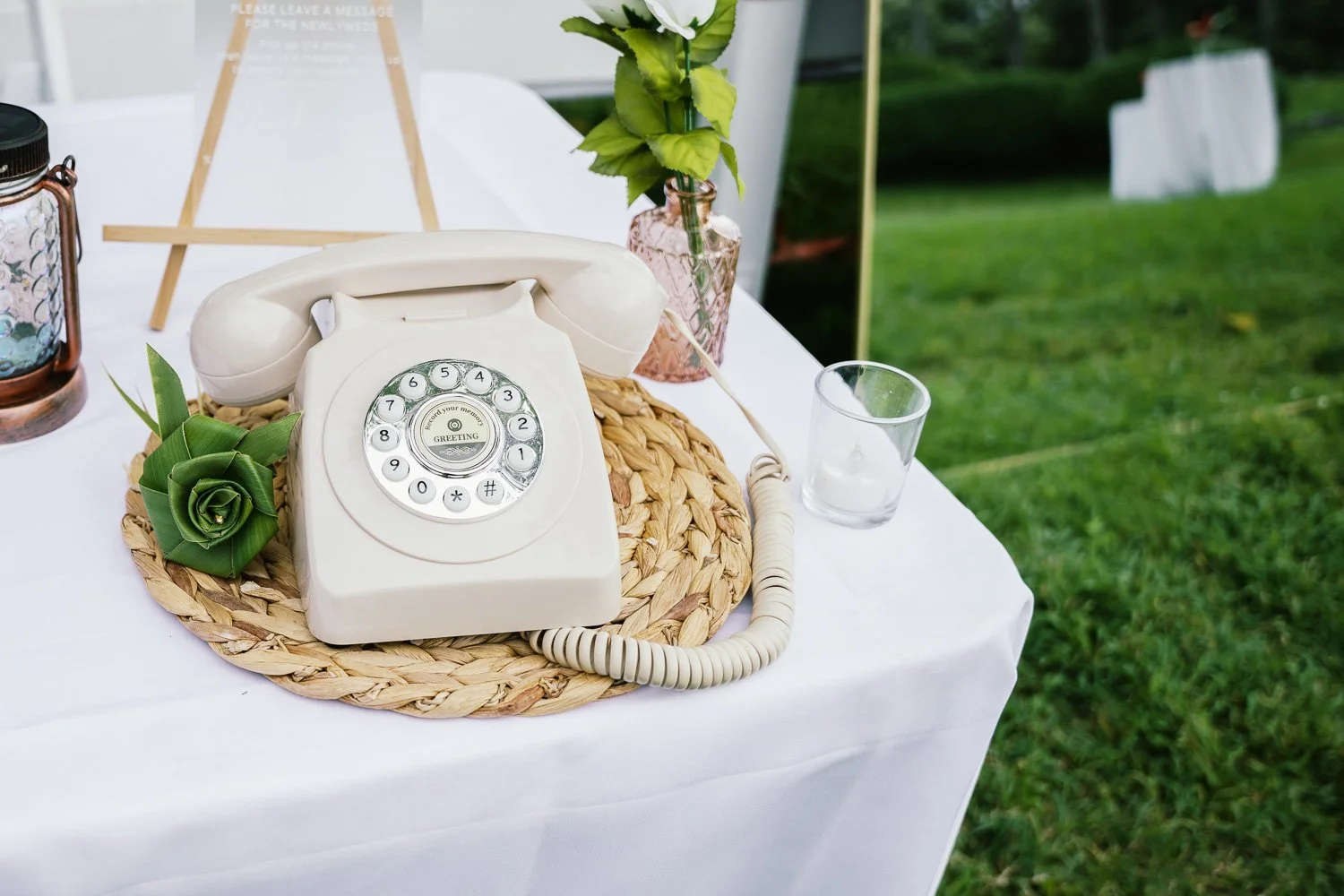 Vintage rotary phone on a table with a woven placemat, decorative flowers, and a glass on a white tablecloth, outdoors with green grass in the background.
