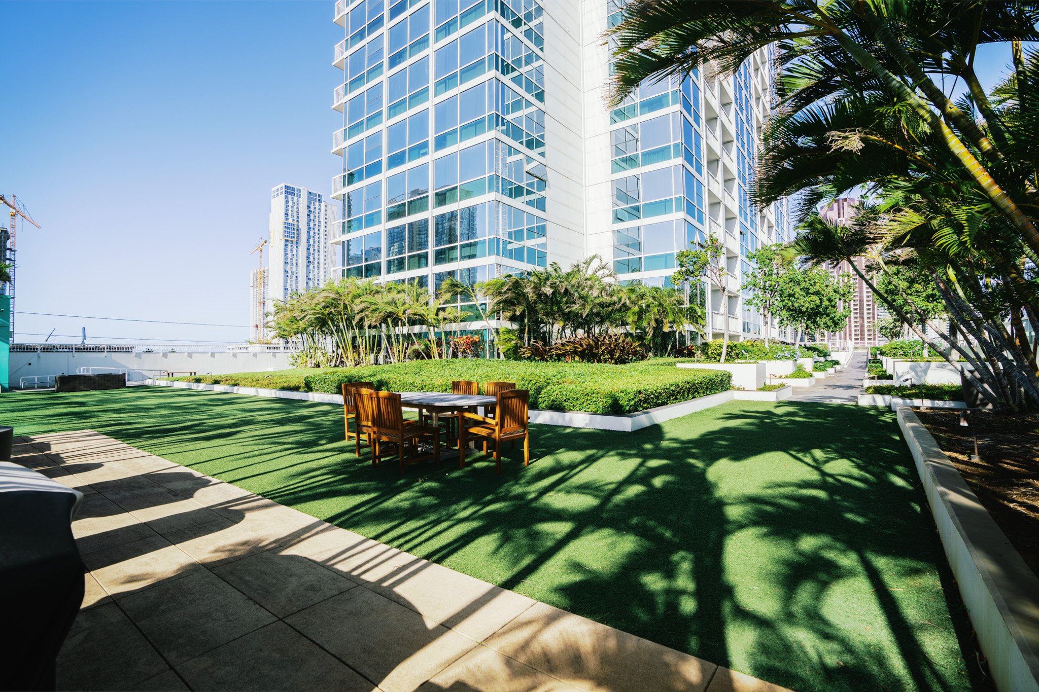 A rooftop garden with green artificial grass, surrounded by lush trees and bushes, and a modern high-rise building with glass windows in the background under a clear blue sky.