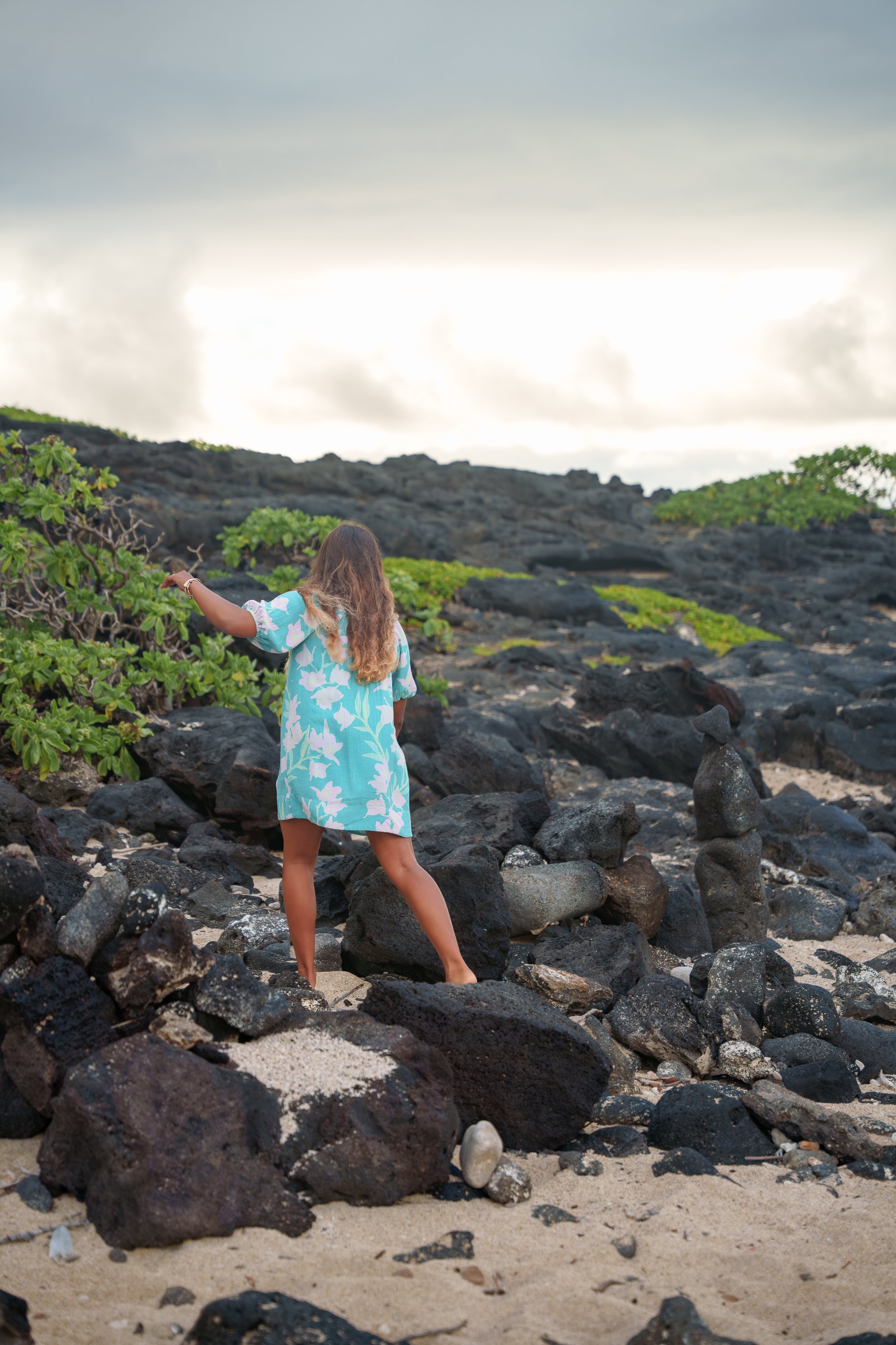 A woman in a light blue and white floral dress standing on a rocky beach, looking at green shrubbery and rocks under a cloudy sky.