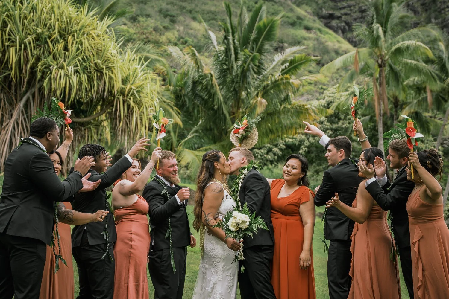 A diverse group of wedding guests celebrating outdoors during a wedding ceremony, with a bride and groom kissing in the center, surrounded by friends holding floral arrangements, lush green tropical plants, and palm trees in the background.