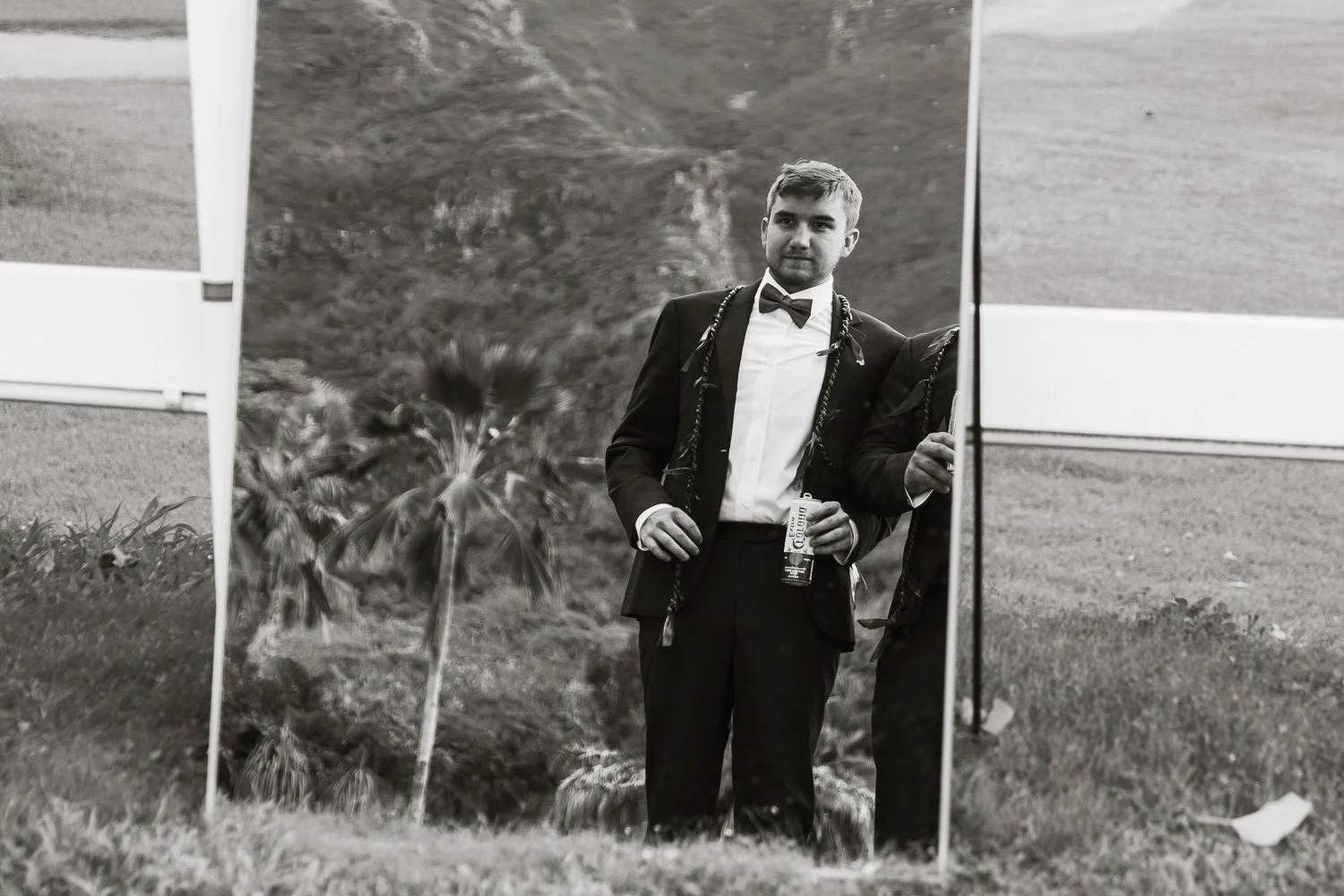 A young man in a tuxedo holding a drink stands outside, reflected in a large mirror or reflective surface, with a hillside and grass in the background.