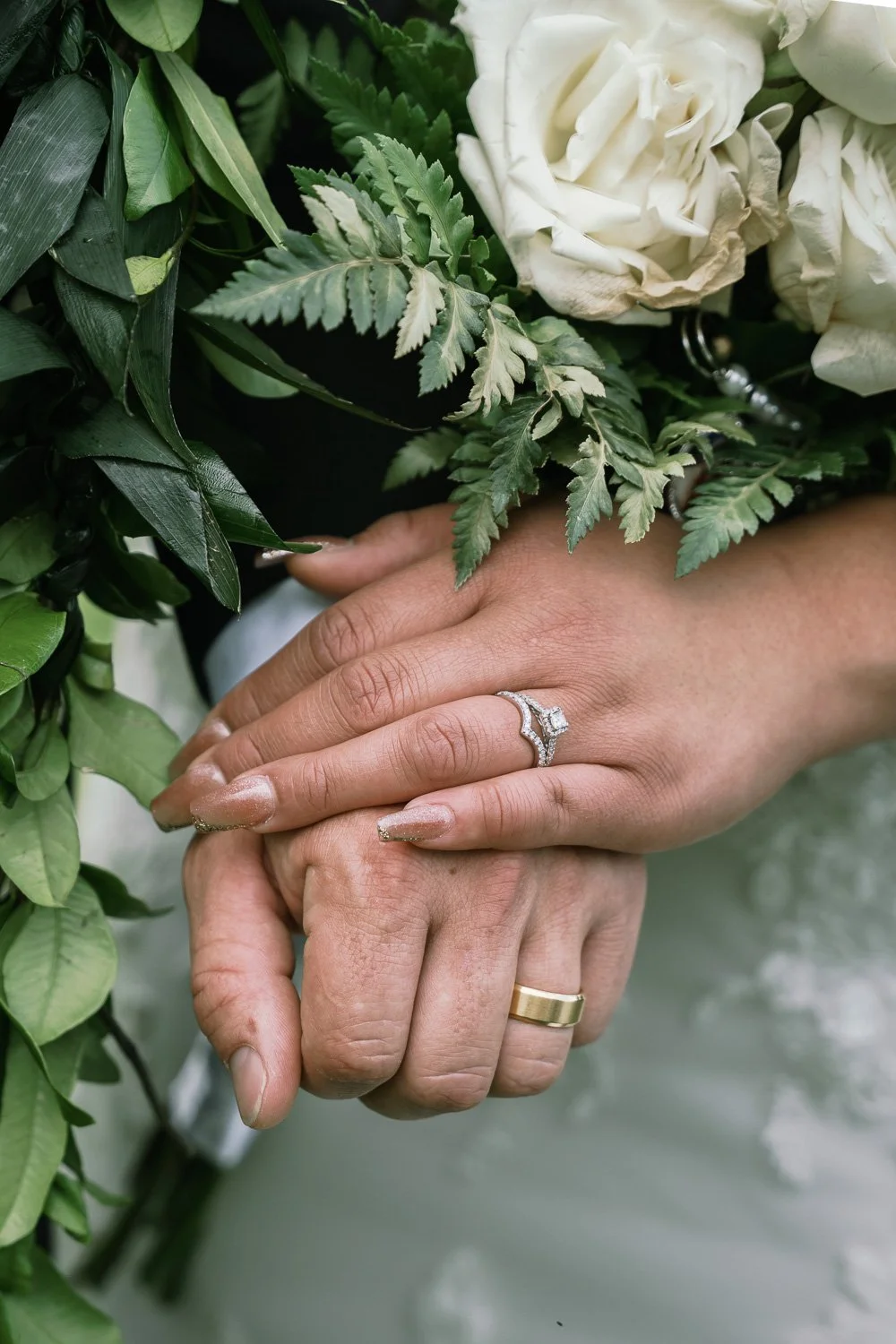 Close-up of two hands with wedding rings, holding each other, surrounded by white roses and green foliage.