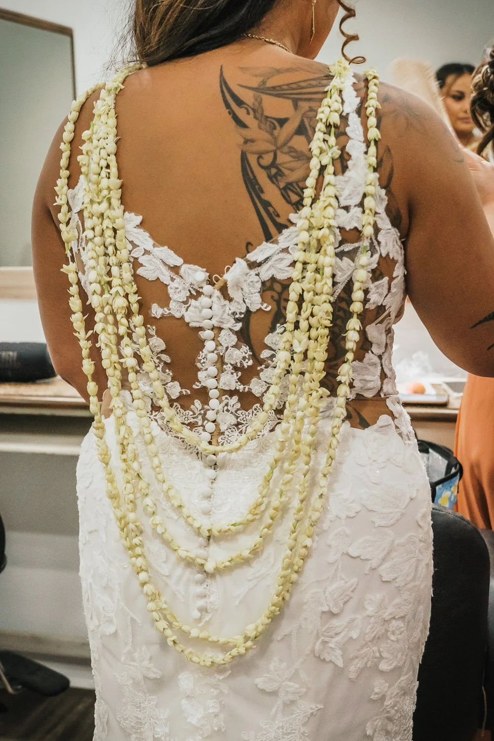 A woman wearing a white lace dress with floral details, adorned with multiple strands of white flowers, standing with her back visible showing a large tattoo on her back and shoulders.