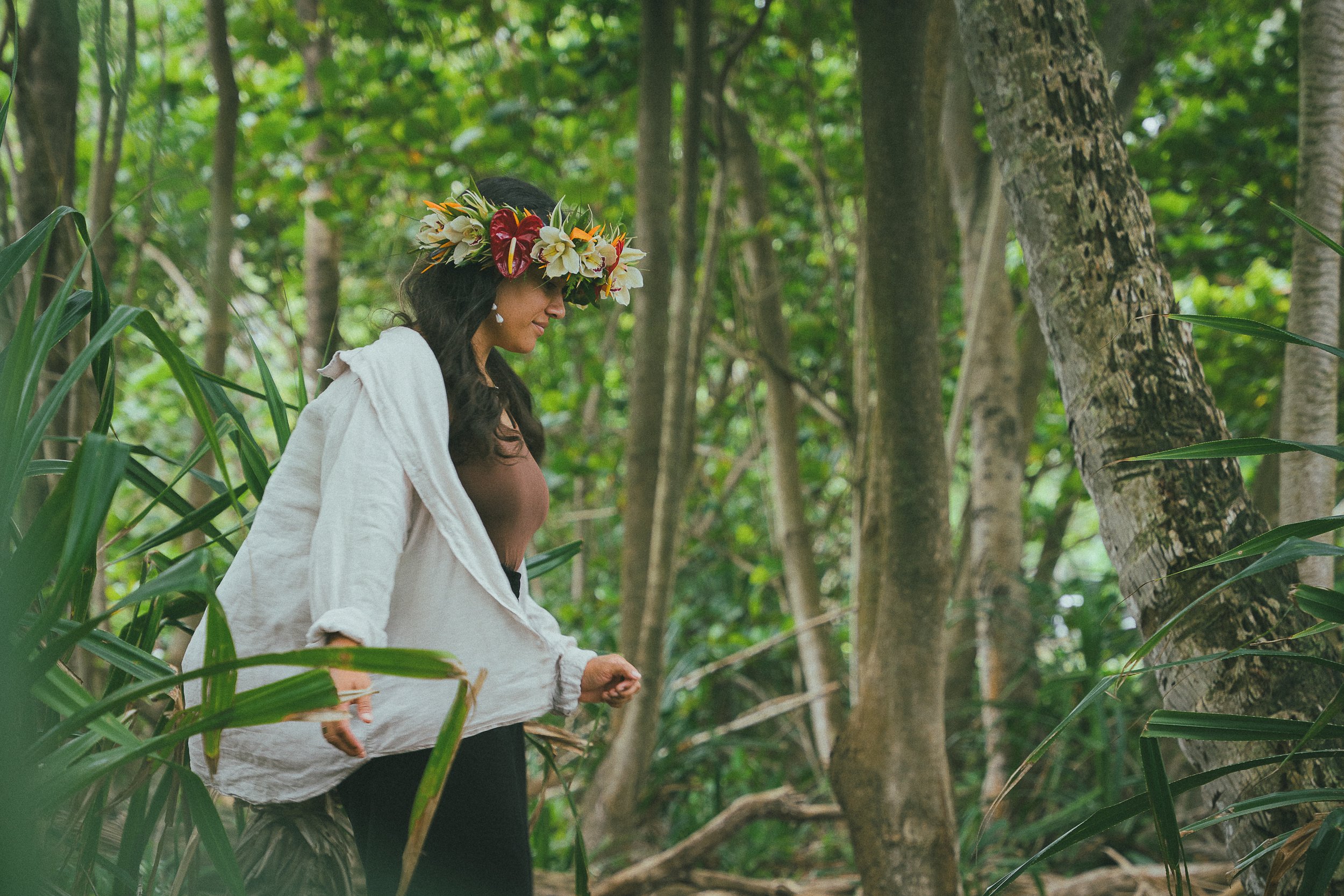 A woman with dark hair wearing a floral crown made of white and red flowers, standing in a lush green forest.
