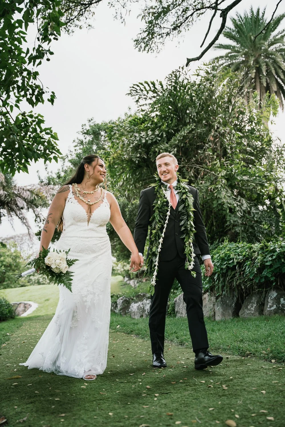 A bride and groom walking hand in hand outdoors during their wedding, surrounded by lush green trees and plants, with the bride holding a bouquet of white flowers.