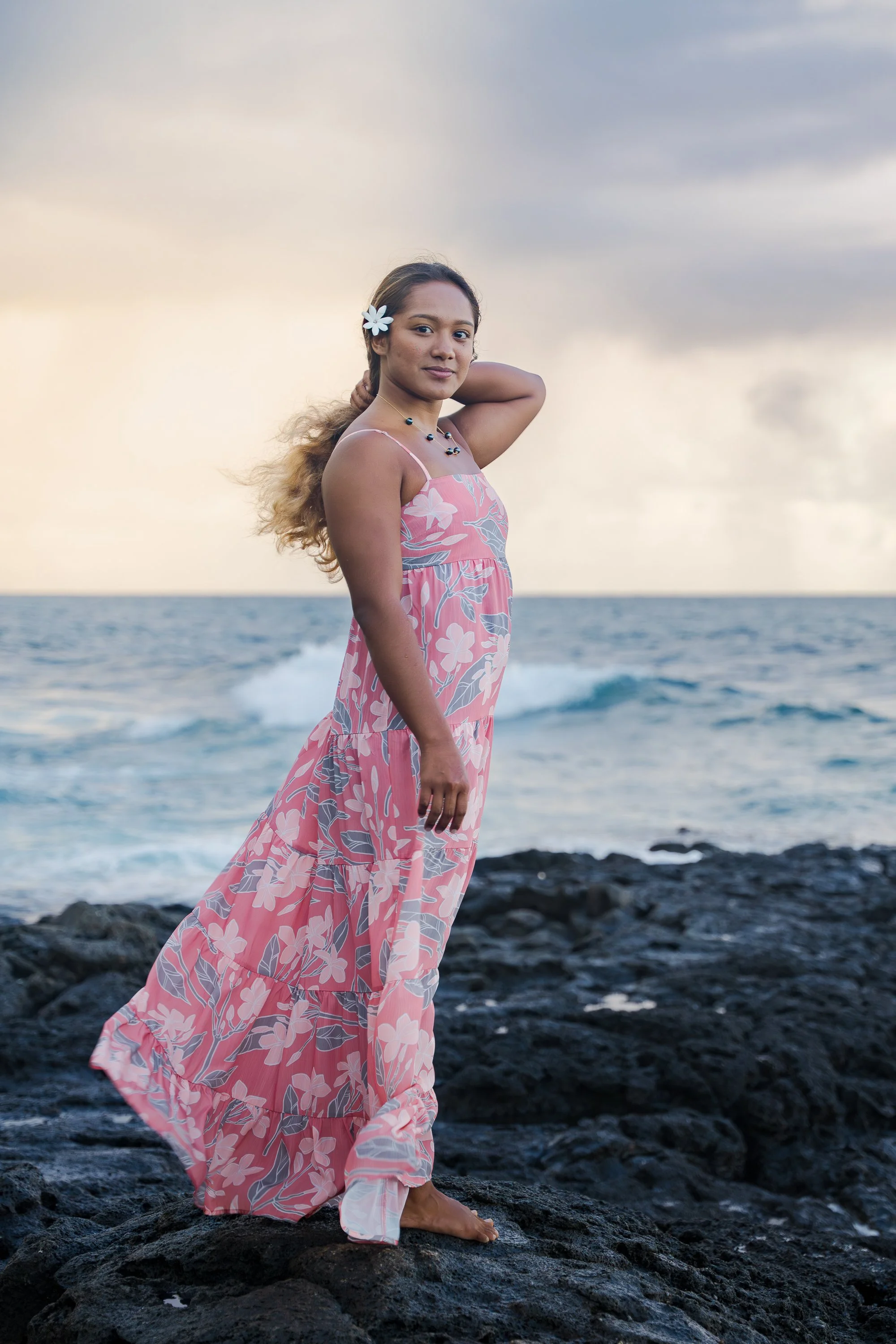 A woman in a pink floral dress standing barefoot on black rocks by the ocean with waves and a cloudy sky in the background.