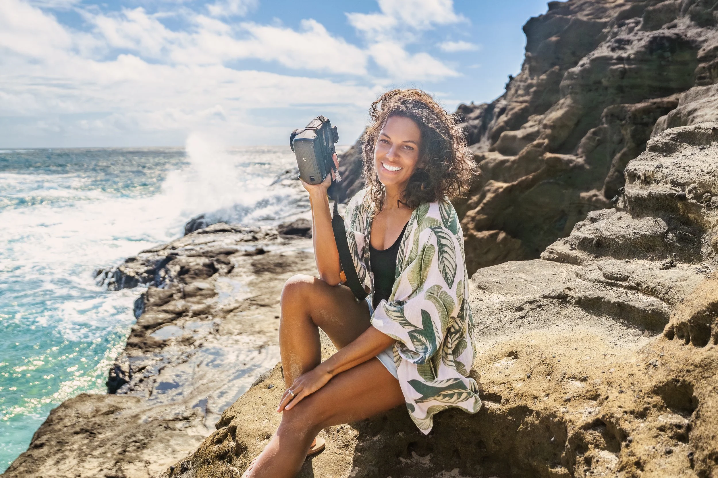 A woman with curly hair sitting on rocks by the ocean, holding a camera, smiling at the camera, wearing a tropical print cover-up and shorts, with waves and a cloudy sky in the background.