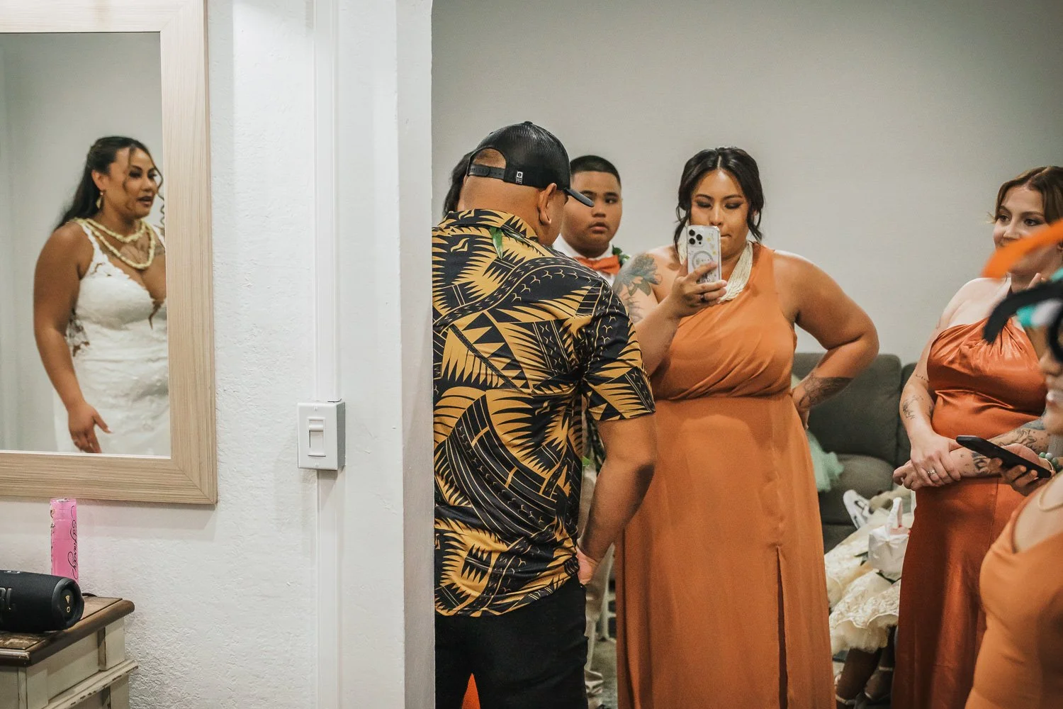 A group of people gathered in a room, with a woman in a white dress reflected in a mirror, and others interacting or looking at their phones.