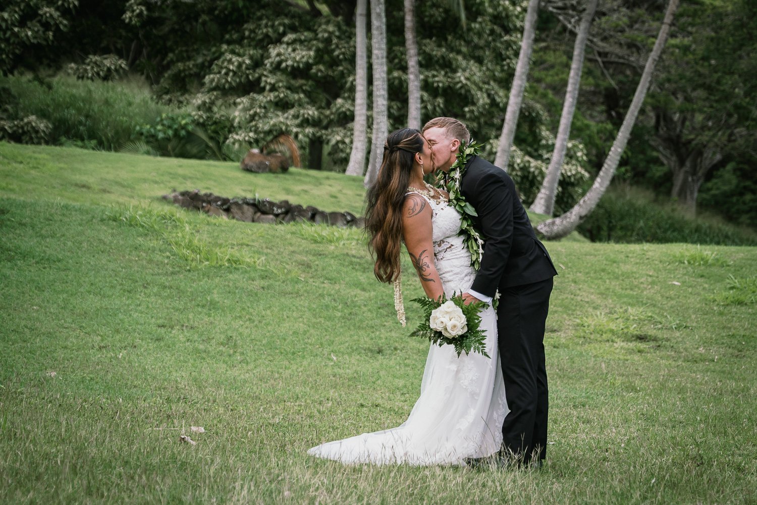 A bride and groom sharing a kiss on a lush green lawn with trees in the background. The bride has long brown hair, tattoos, and is holding a bouquet of white flowers. The groom is in a black suit with a green leafy lei. They are outdoors in a natural