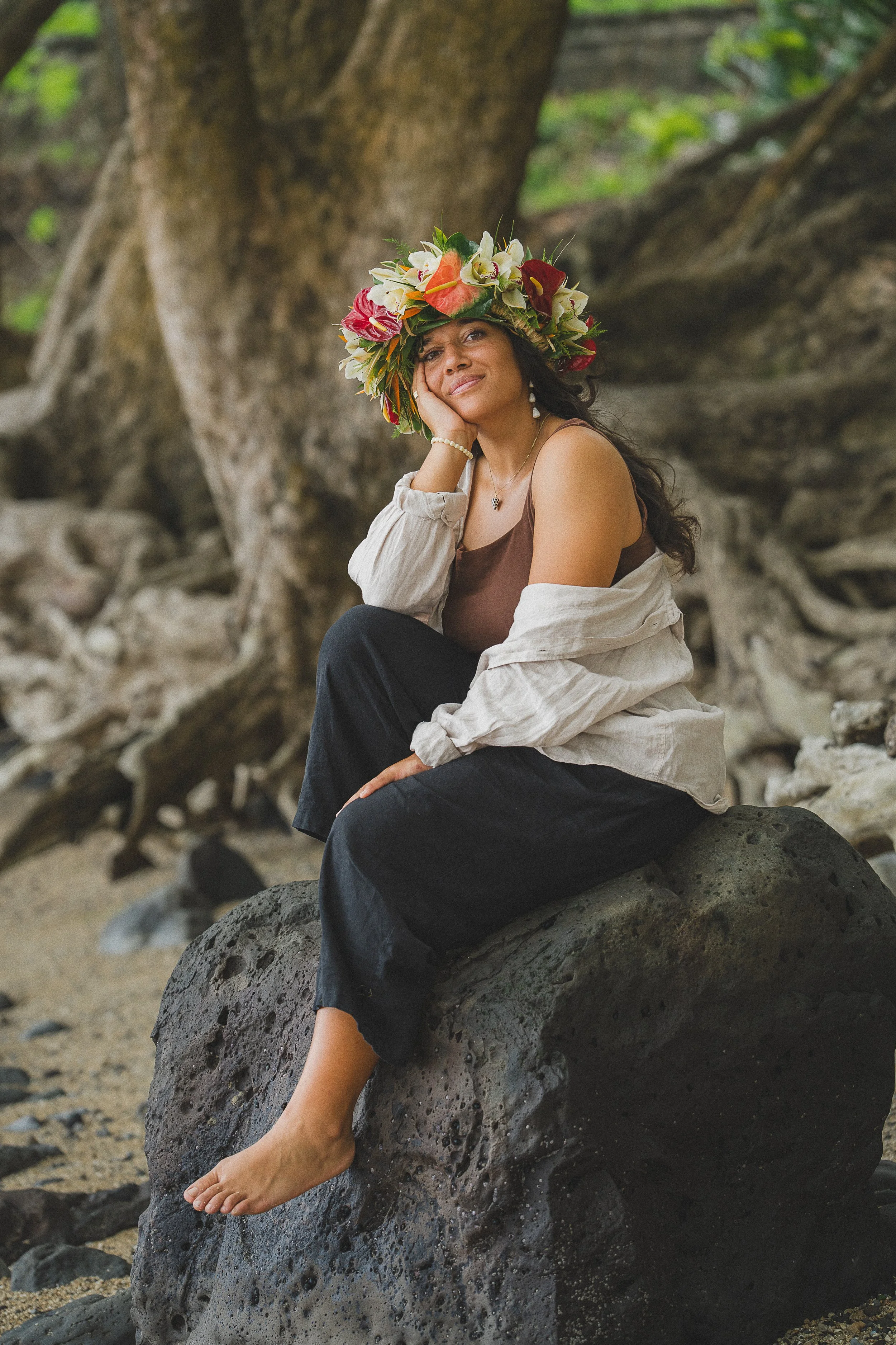 A woman sitting barefoot on a large volcanic rock on a beach, wearing a floral crown, a brown tank top, and a light-colored shirt. She has long dark hair and is resting her chin on her hand, looking at the camera with a slight smile.