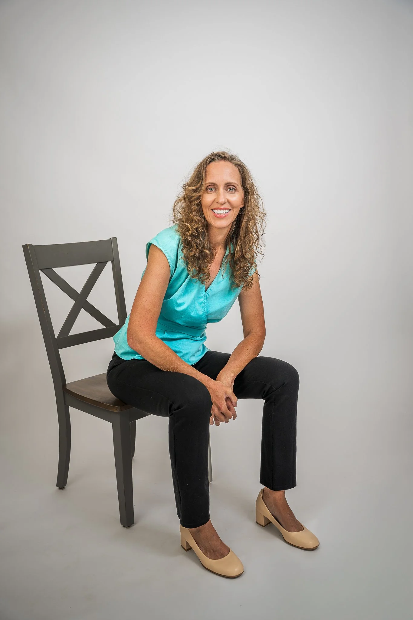 A woman with curly brown hair, wearing a turquoise sleeveless top and black pants, sitting on a dark gray chair with crossed back, smiling at the camera against a plain white background.