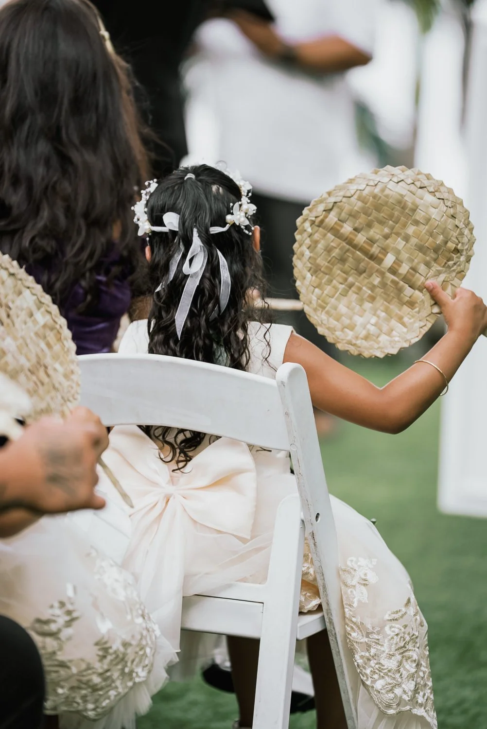 Child wearing a white dress with lace details, sitting on a white chair, holding a woven fan, attending a celebration or ceremony.