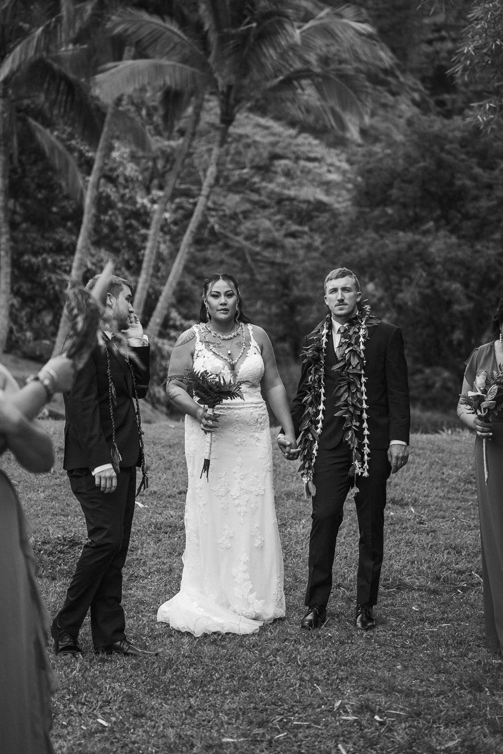 A black and white photo of a couple holding hands during their wedding ceremony outdoors, with palm trees and lush greenery in the background, and wedding attendants surrounding them.