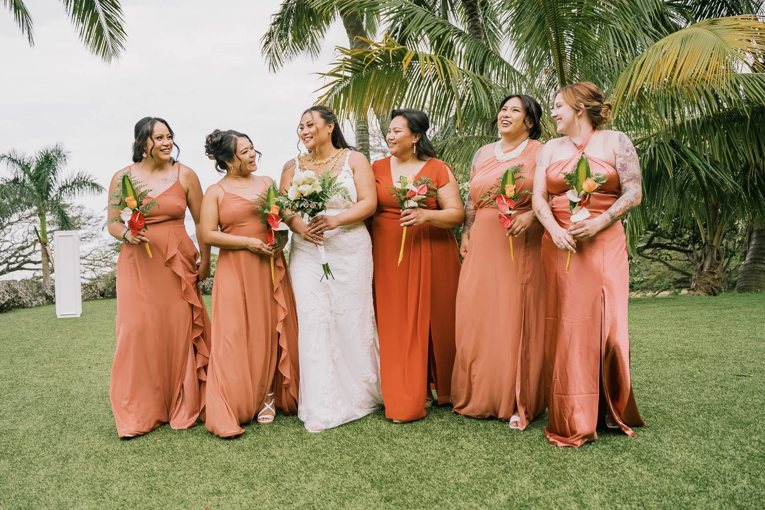 A bride and five bridesmaids standing on a grassy lawn with palm trees in the background, all holding small bouquets and smiling together.