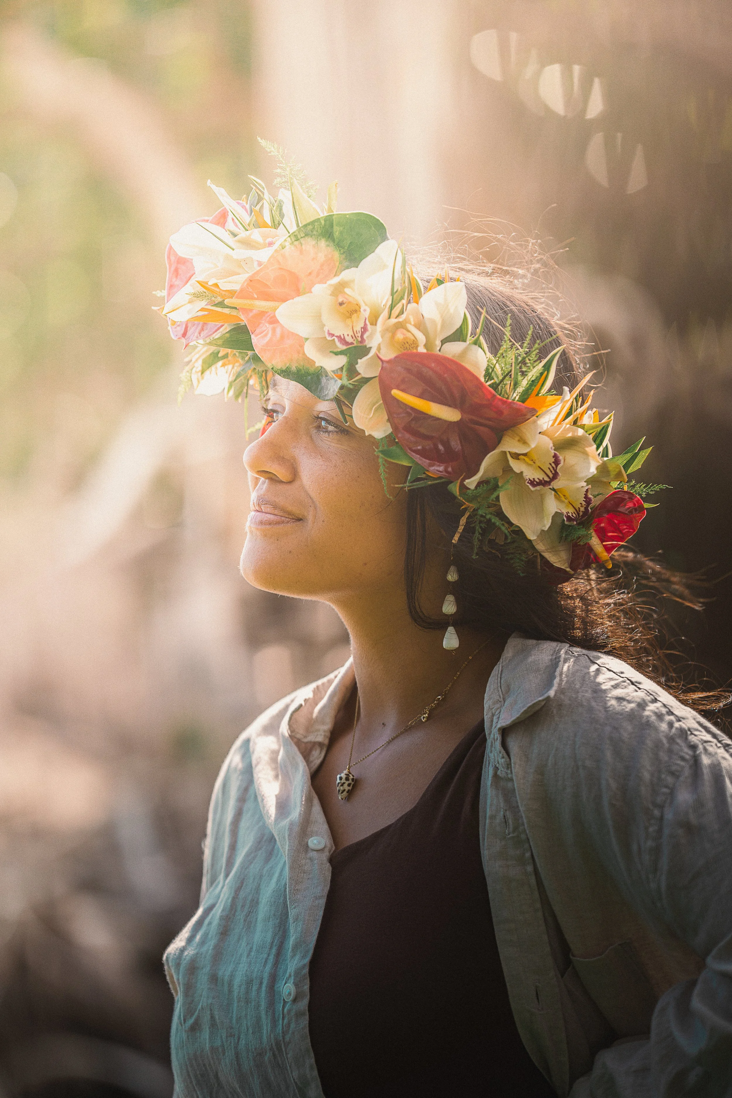 A woman wearing a floral crown with white, pink, orange, and red flowers, gazing thoughtfully into the distance outside.