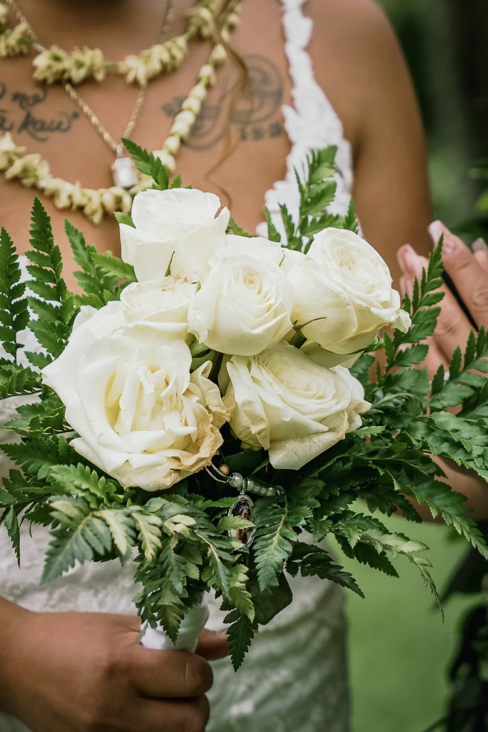 Close-up of a woman holding a bridal bouquet of white roses and green ferns, with tattoos visible on her chest and shoulder, wearing lace straps and a floral necklace.