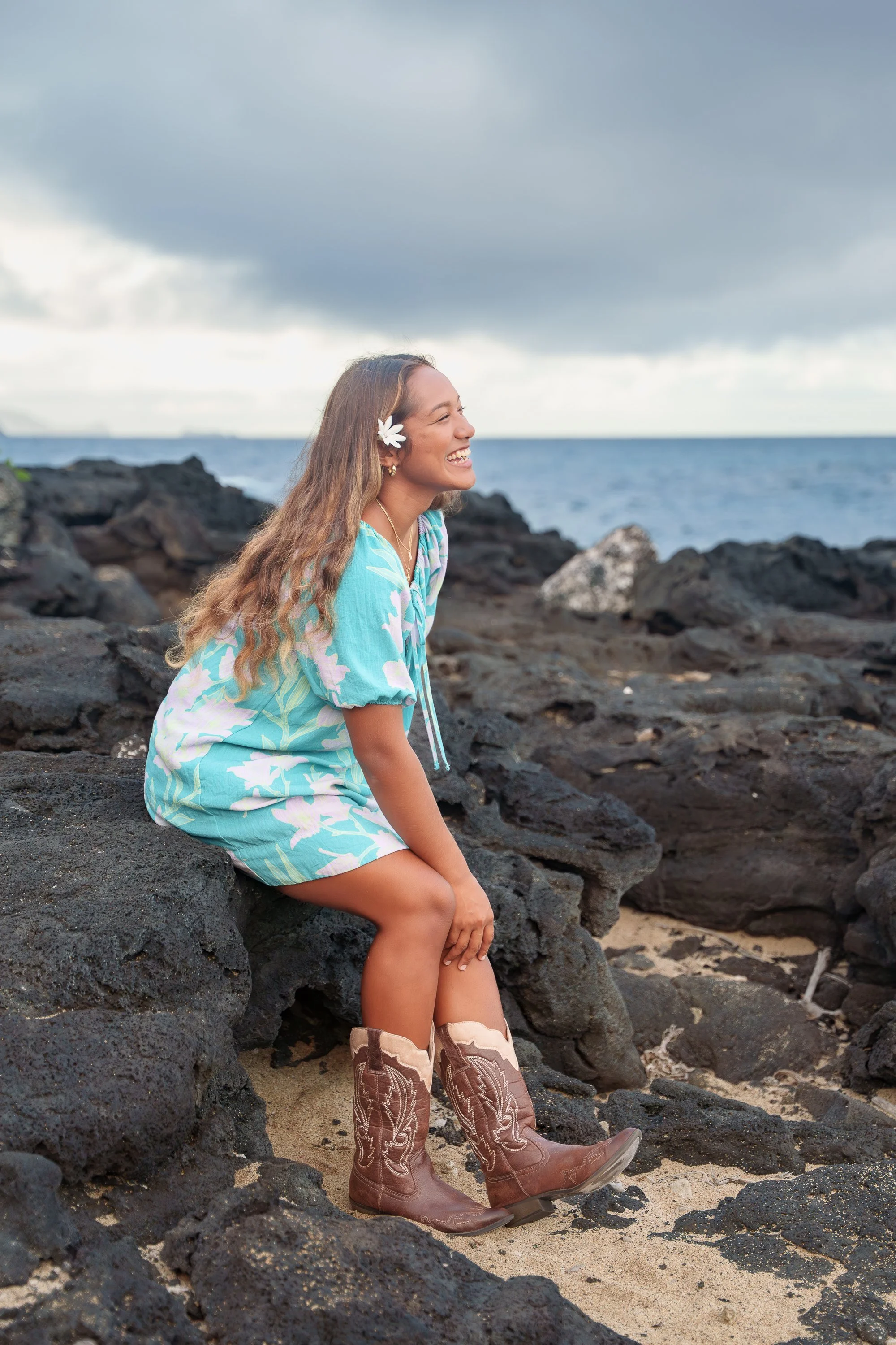 A woman with long wavy hair wearing a blue floral dress, a white flower in her hair, and cowboy boots sitting on rocks by the beach, smiling and looking to her right with the ocean and cloudy sky in the background.