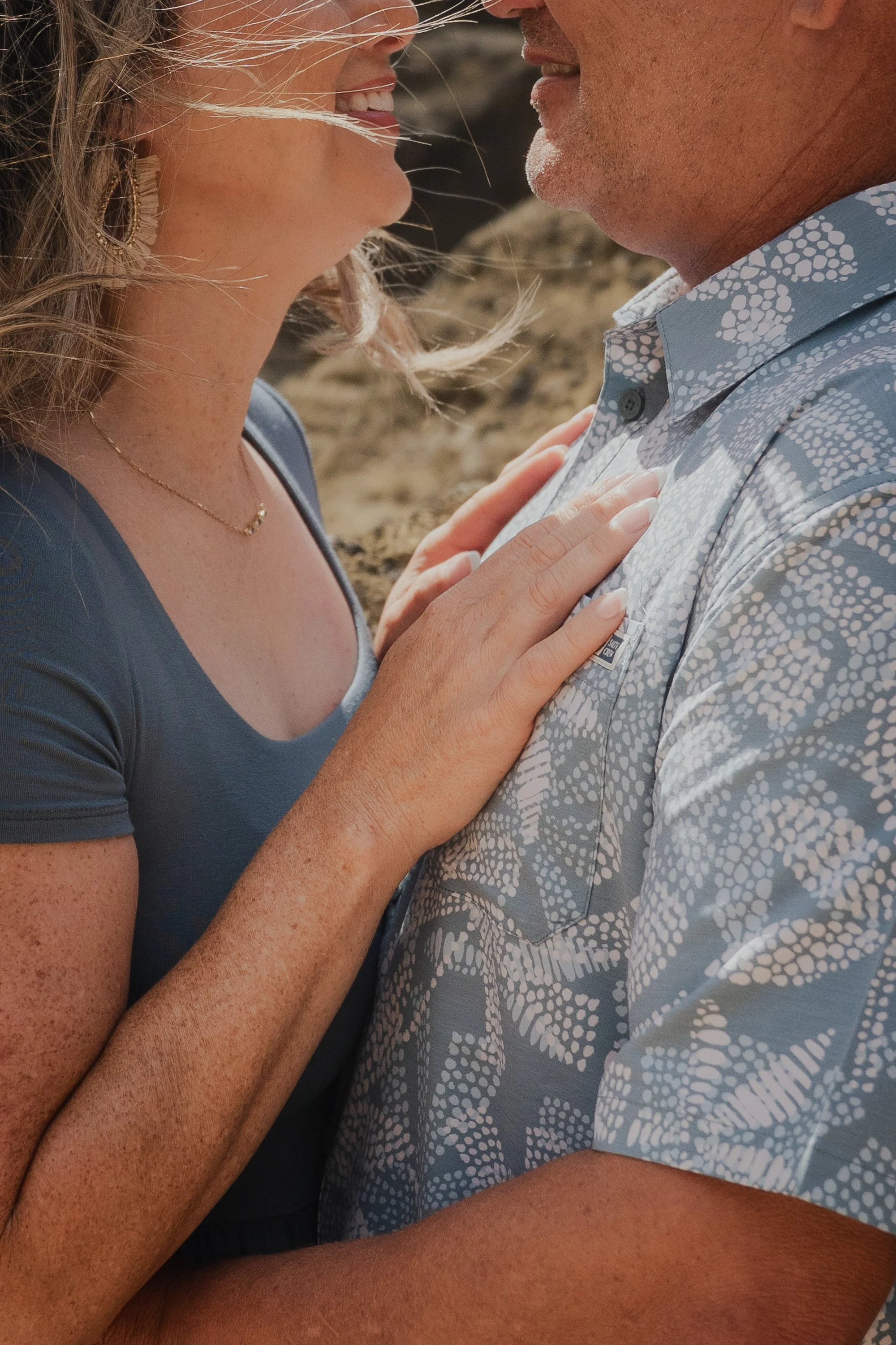 A couple standing close together, smiling, with the woman's hand on the man's chest, on a sandy beach.