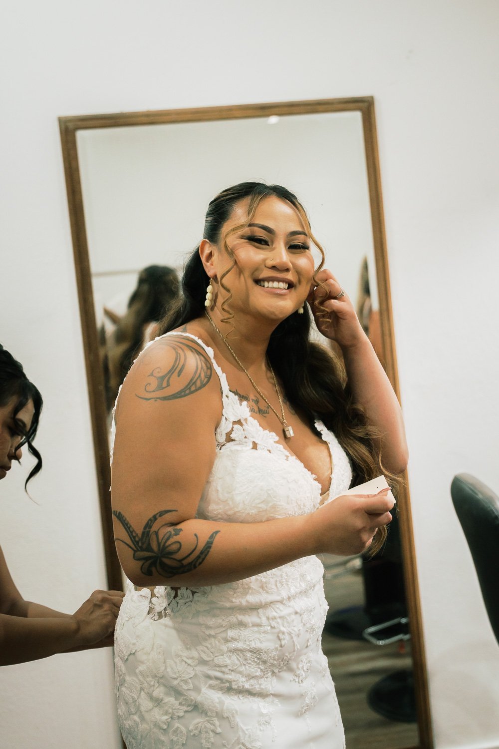 Woman in a white lace dress smiling and touching her hair, tattoos on her arm, standing in front of a mirror.