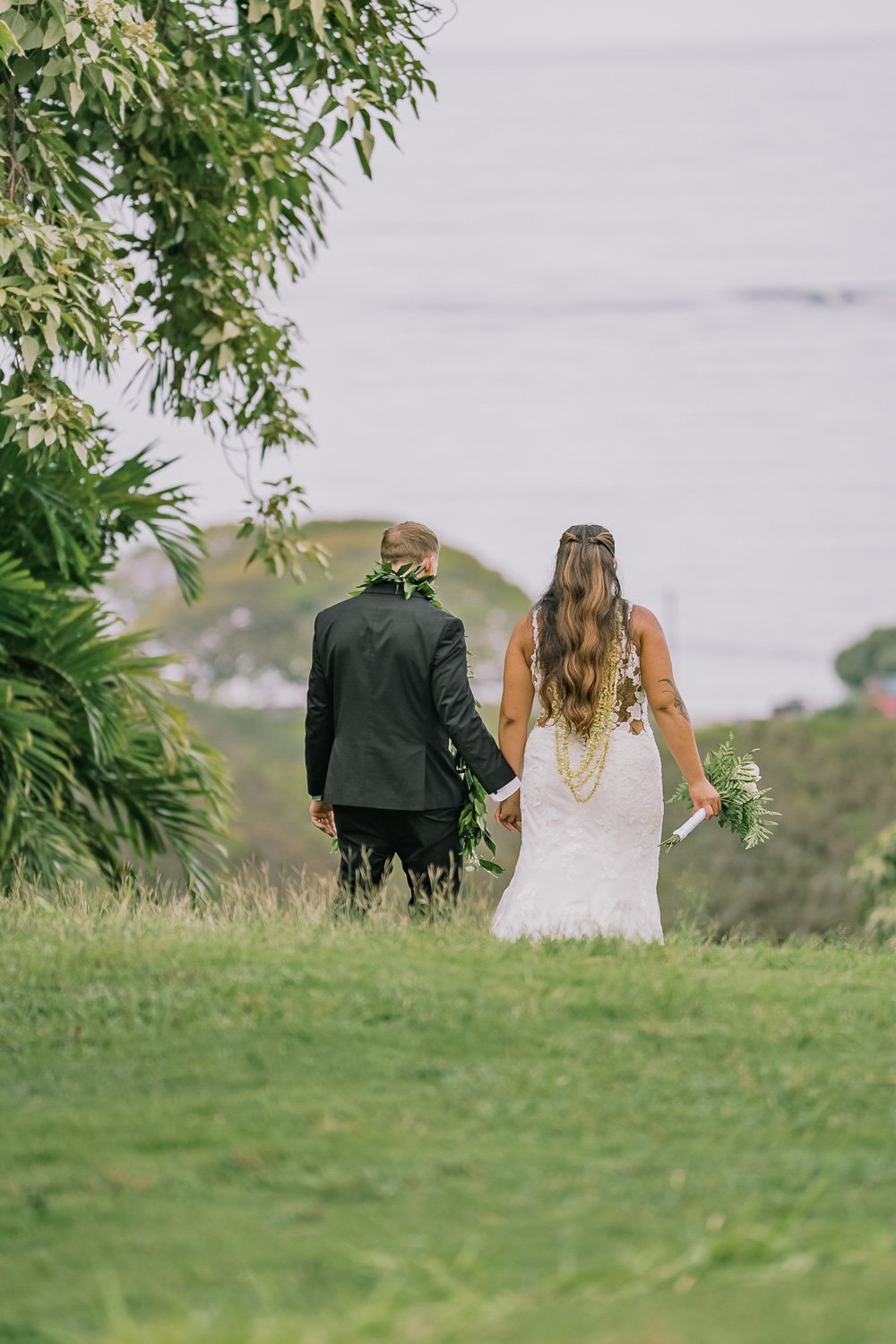 A bride and groom walk hand in hand outdoors near a body of water, with lush greenery framing the scene.