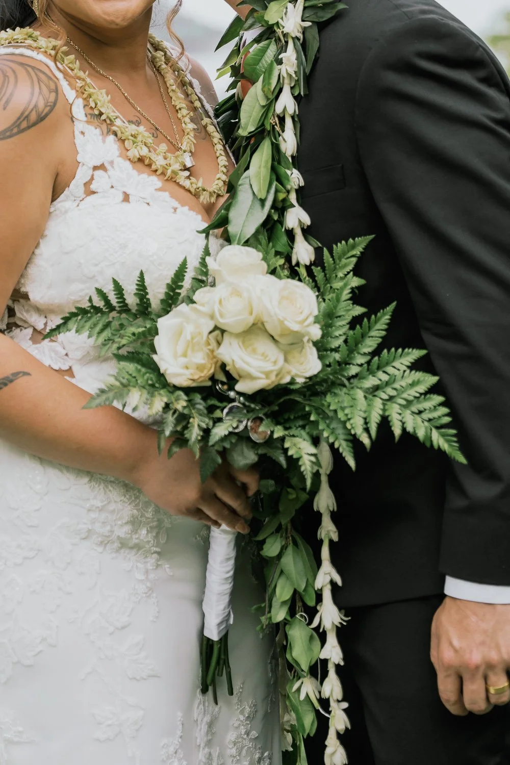Close-up of a bride and groom holding a wedding bouquet, featuring white roses and green foliage, with bride wearing a white lace dress and various necklaces, and groom in a black suit with a floral boutonniere.