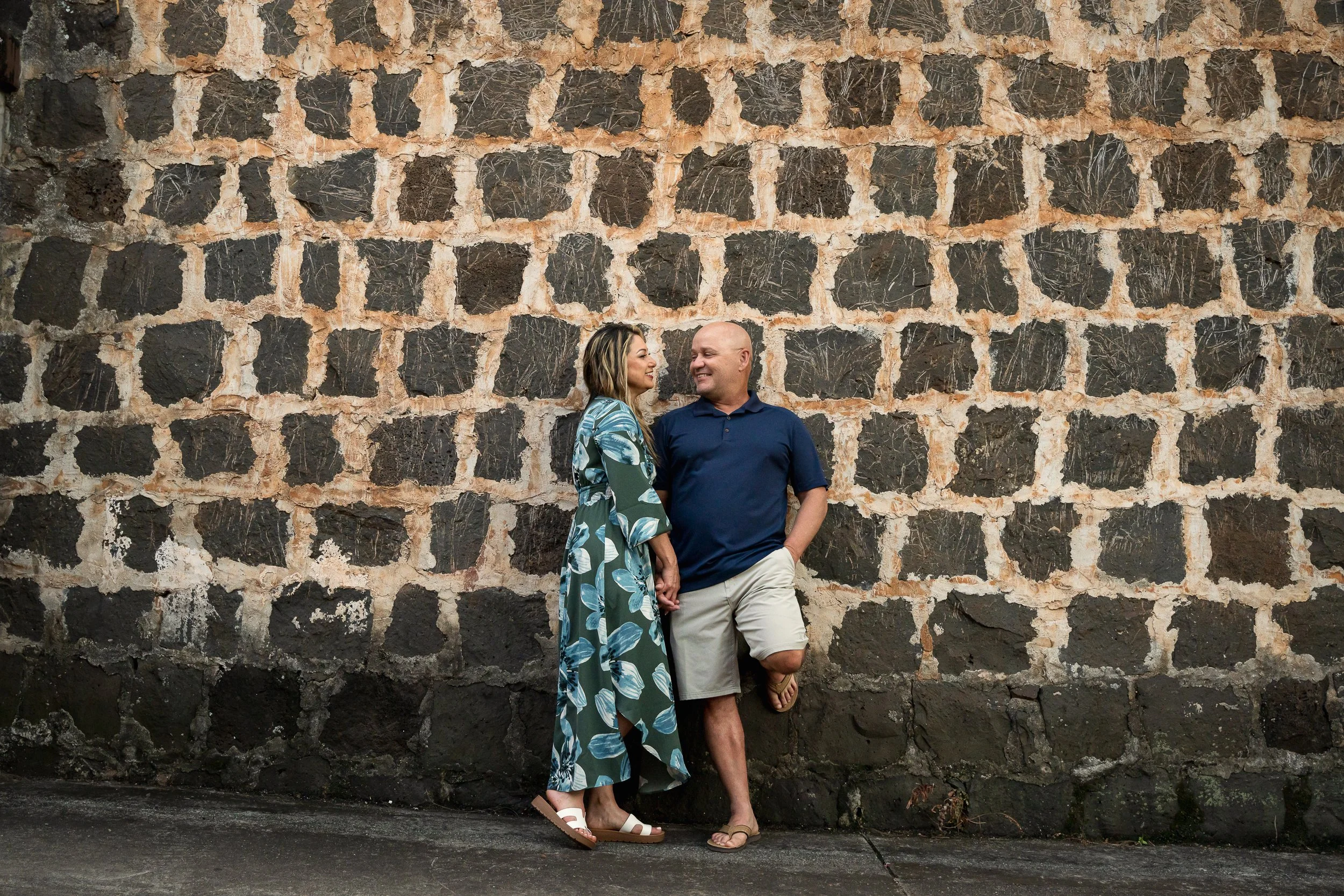 A man and woman standing against a large, rustic stone wall. The woman is wearing a long, patterned dress and sandals, while the man is dressed in a navy polo shirt and kh shorts, both smiling and looking at each other.