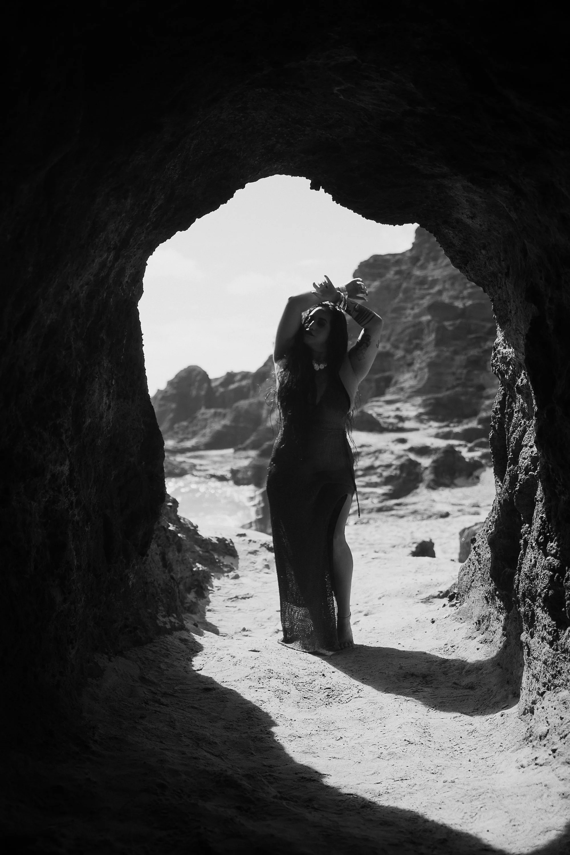 Black and white photo of a woman standing in a cave opening on a beach, with rocky cliffs in the background, wearing a long dress and jewelry, posing with her arms above her head.
