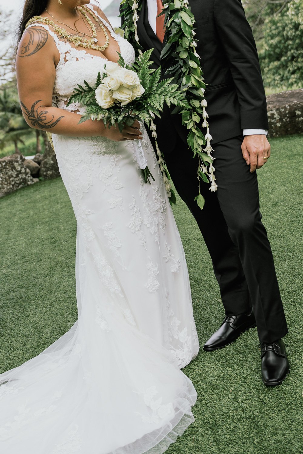 Close-up of a bride and groom at their wedding, the bride holding a bouquet of white roses and greenery, wearing a lace wedding dress with tattoos on her arm, and the groom in a black suit with a floral lei, outdoors on green grass.