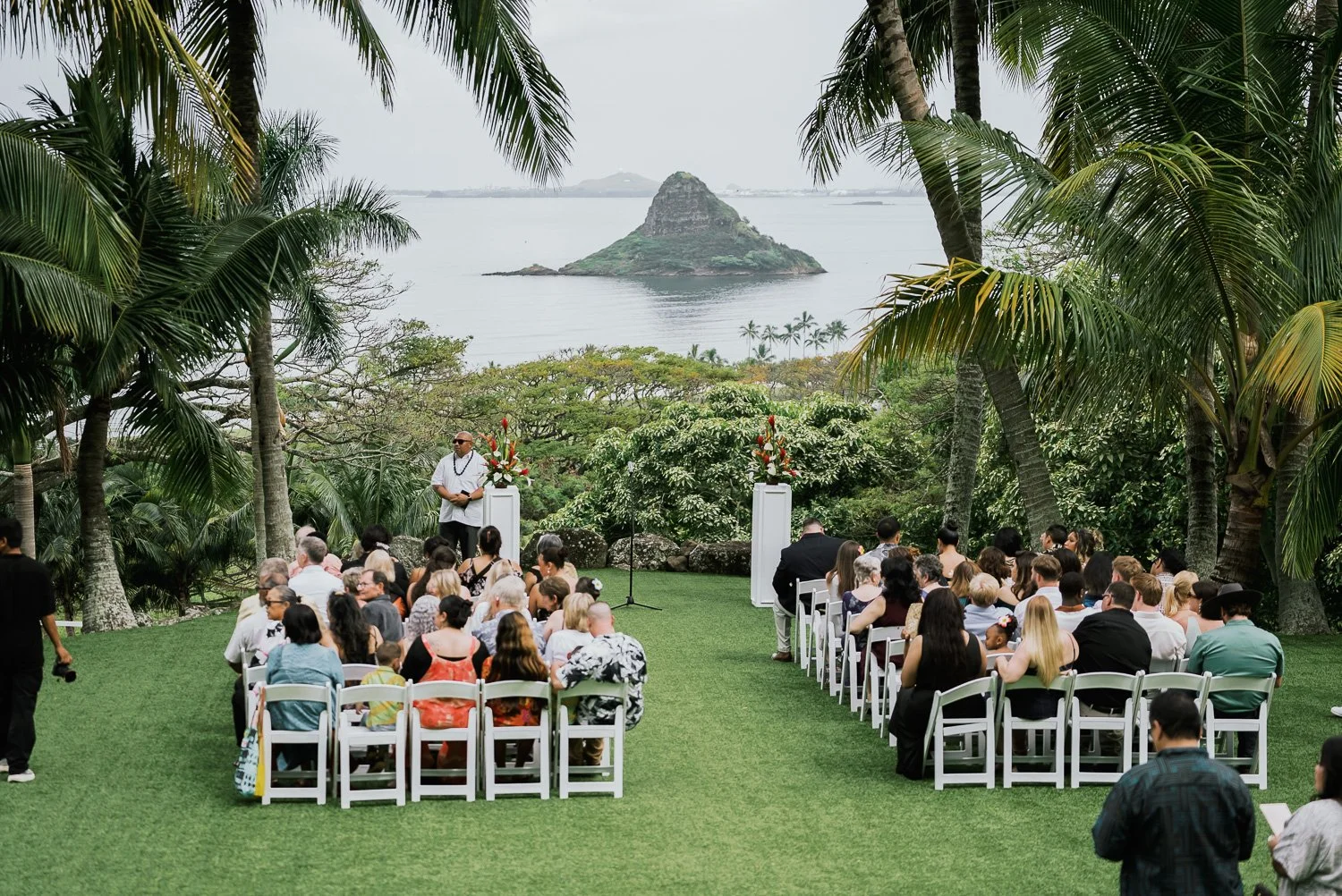 Outdoor wedding ceremony with seated guests and ocean view of a small island, surrounded by palm trees.