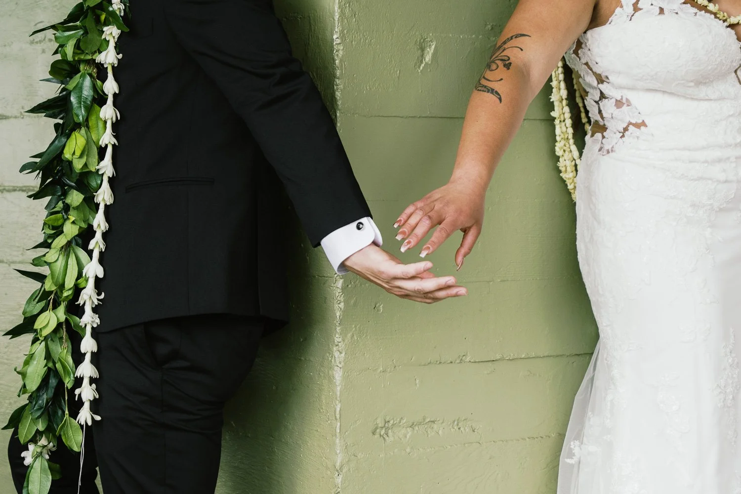 A bride and groom touching hands with fingers close, standing against a green wall during a wedding ceremony.