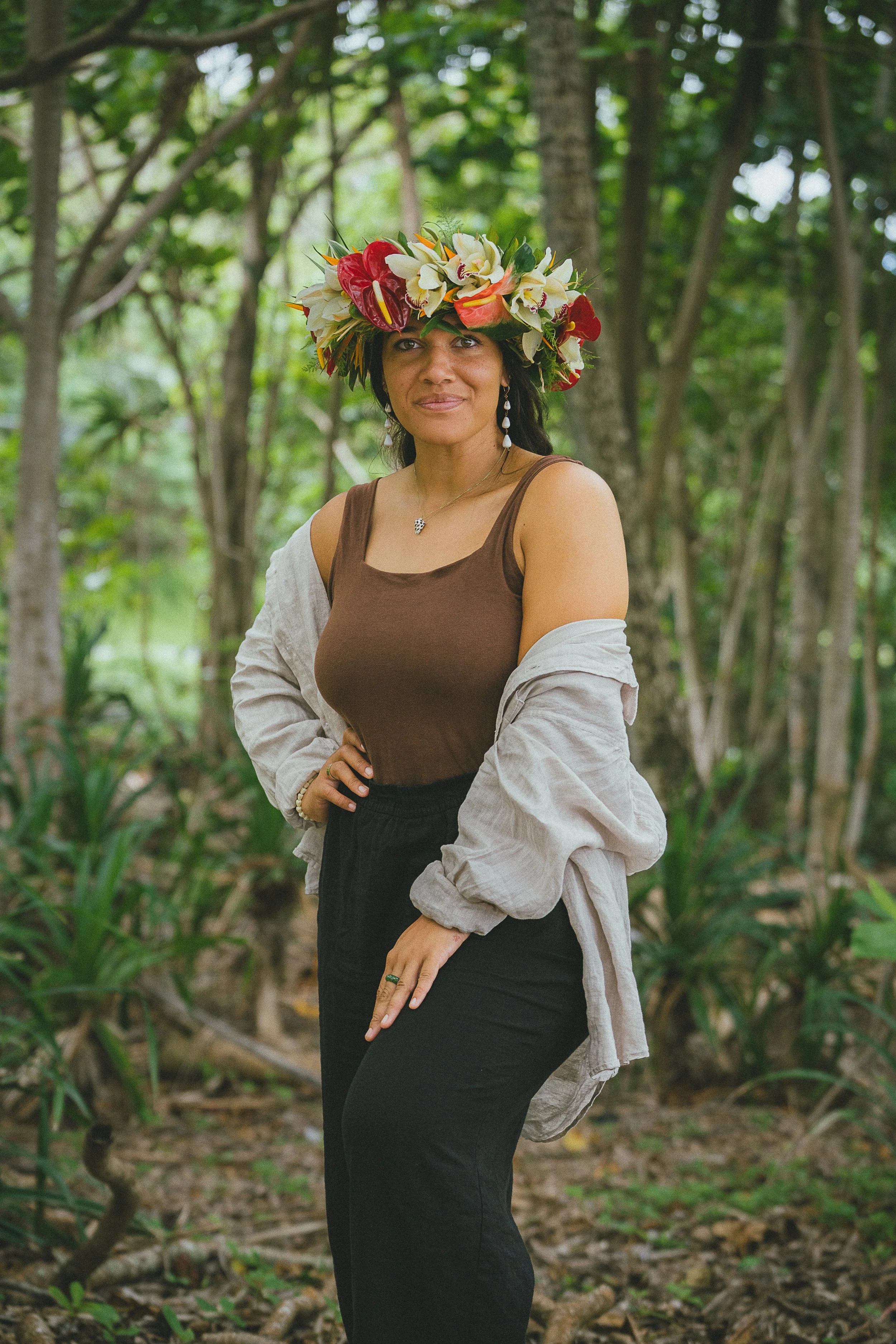 A woman with medium skin tone wearing a floral crown made of orchids, red anthuriums, and other flowers, standing outdoors in a lush green forest.