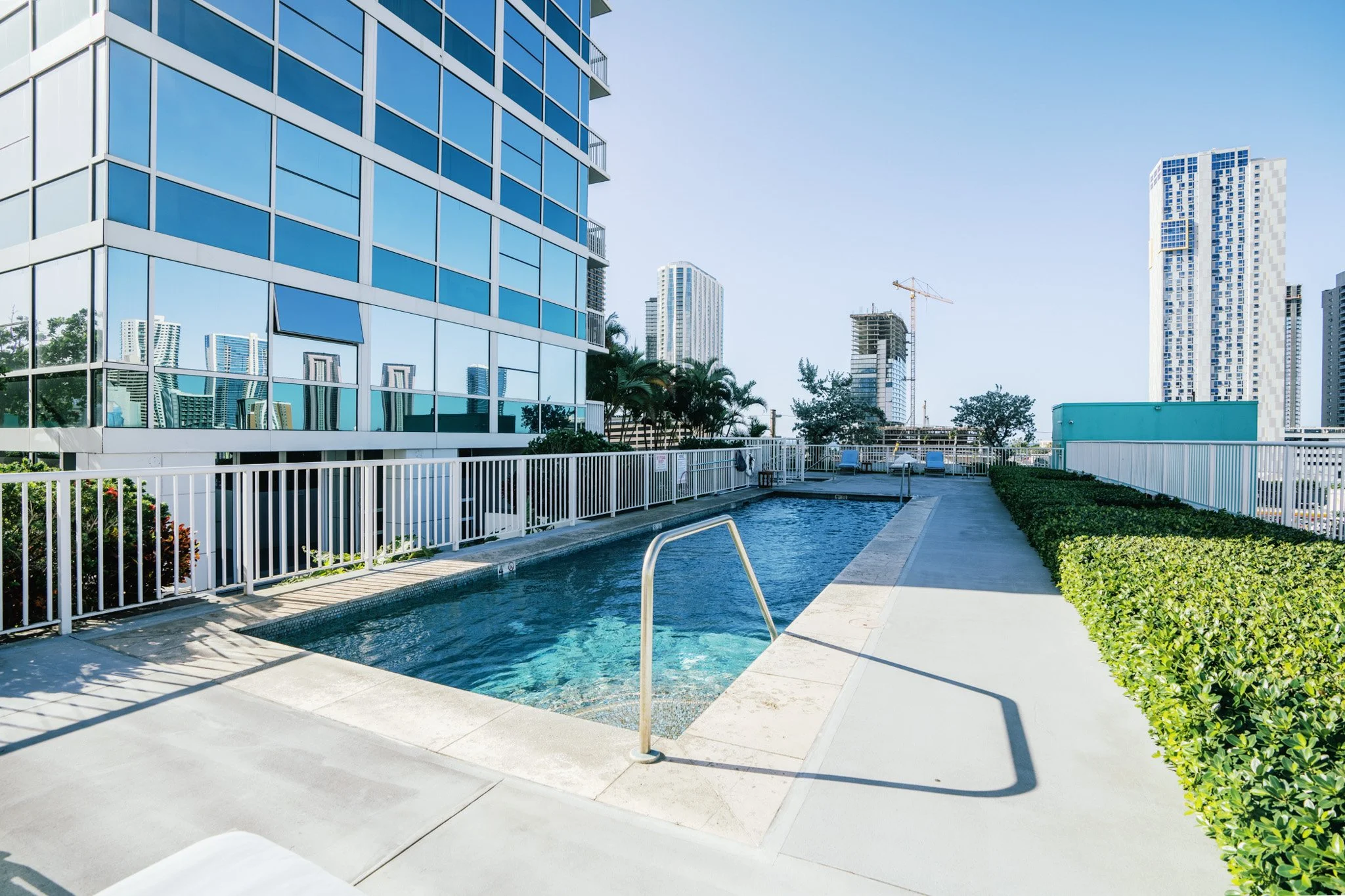 An outdoor pool area on a building rooftop with a city skyline in the background. The pool is surrounded by a white fence, with lounge chairs and greenery nearby. Tall modern buildings and construction cranes are visible in the distance, set against a clear blue sky.