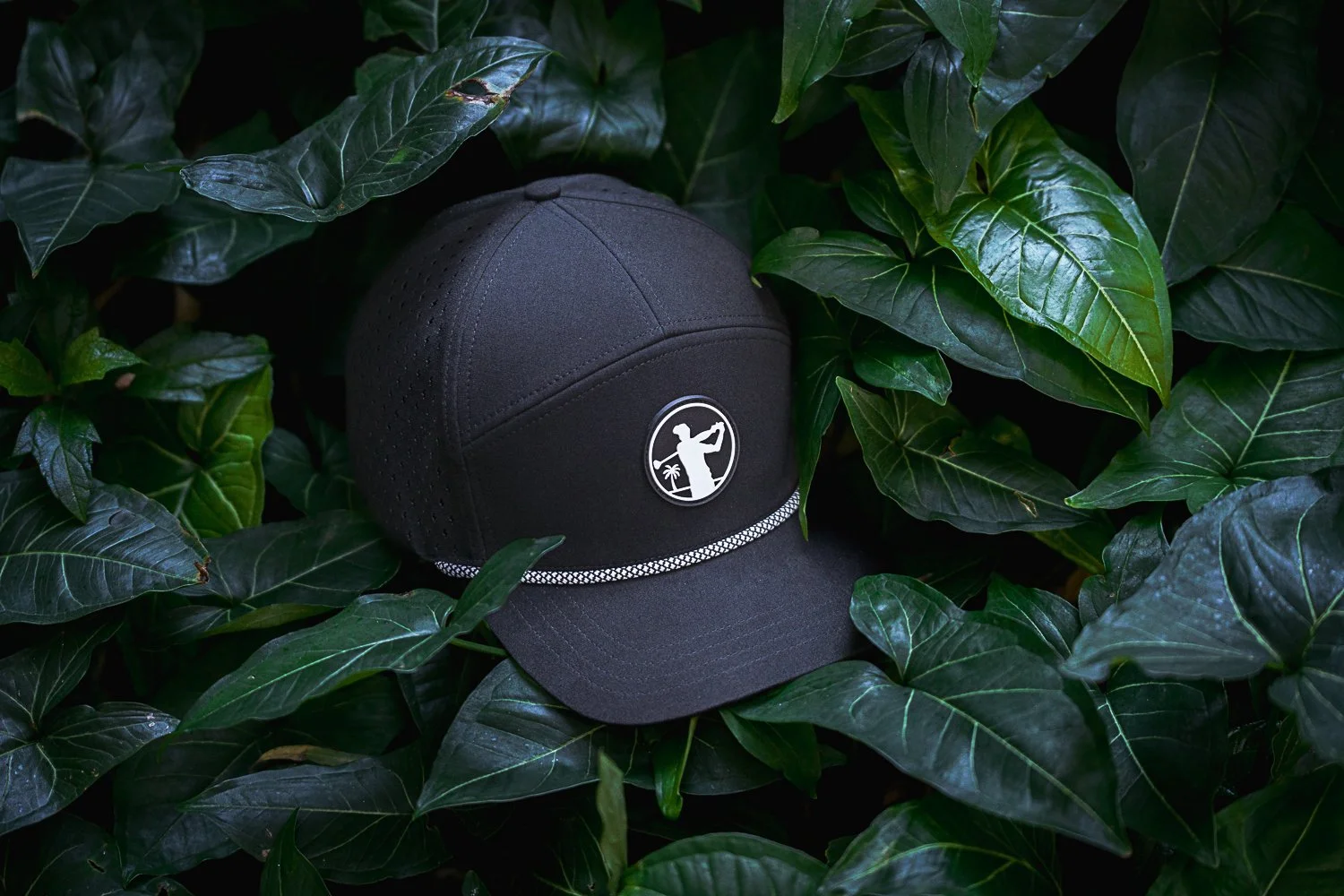 A black baseball cap with a round logo featuring a golf figure and a palm tree, surrounded by lush green leaves.