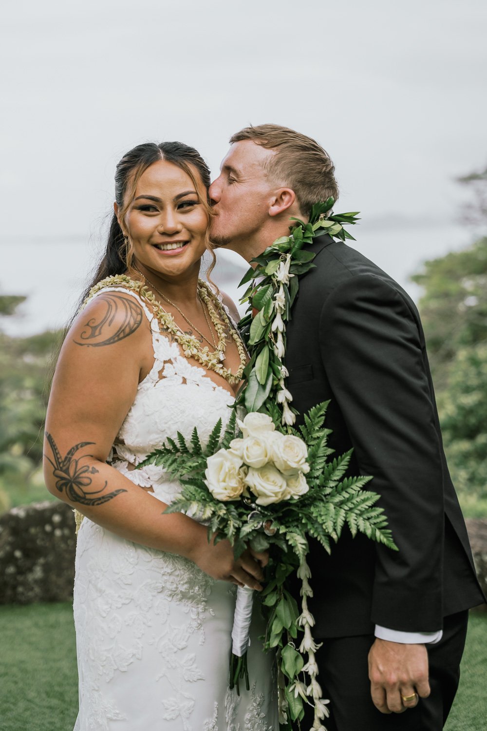A newlywed couple, with the groom kissing the bride on the cheek, both dressed in wedding attire outdoors during daytime. The bride is holding a bouquet of white roses and greenery, and has tattoos on her arms.