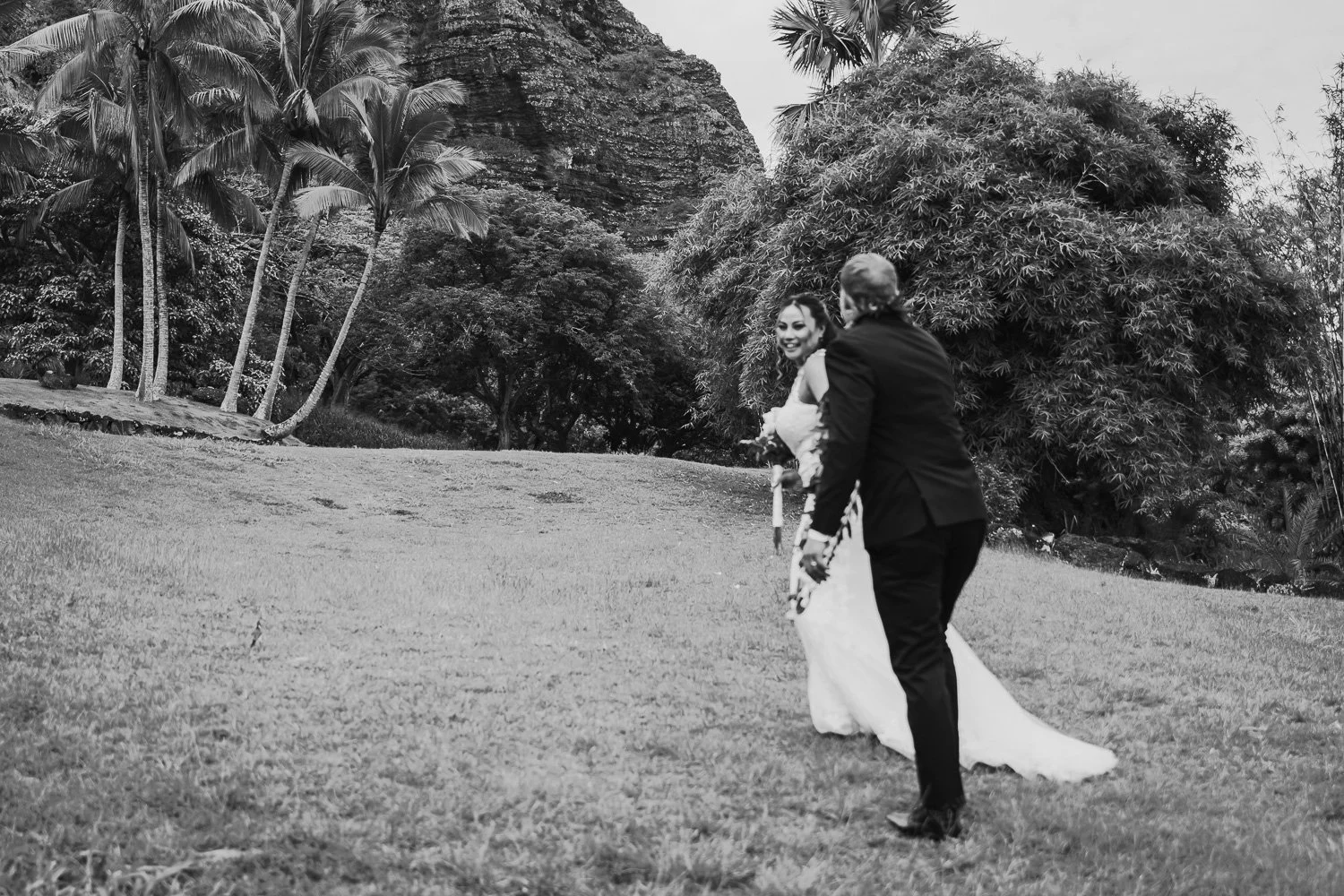 A black and white photograph of a wedding couple outdoors, with the woman in a wedding dress and the man in a suit. They are on a grassy area, surrounded by trees and tropical plants.