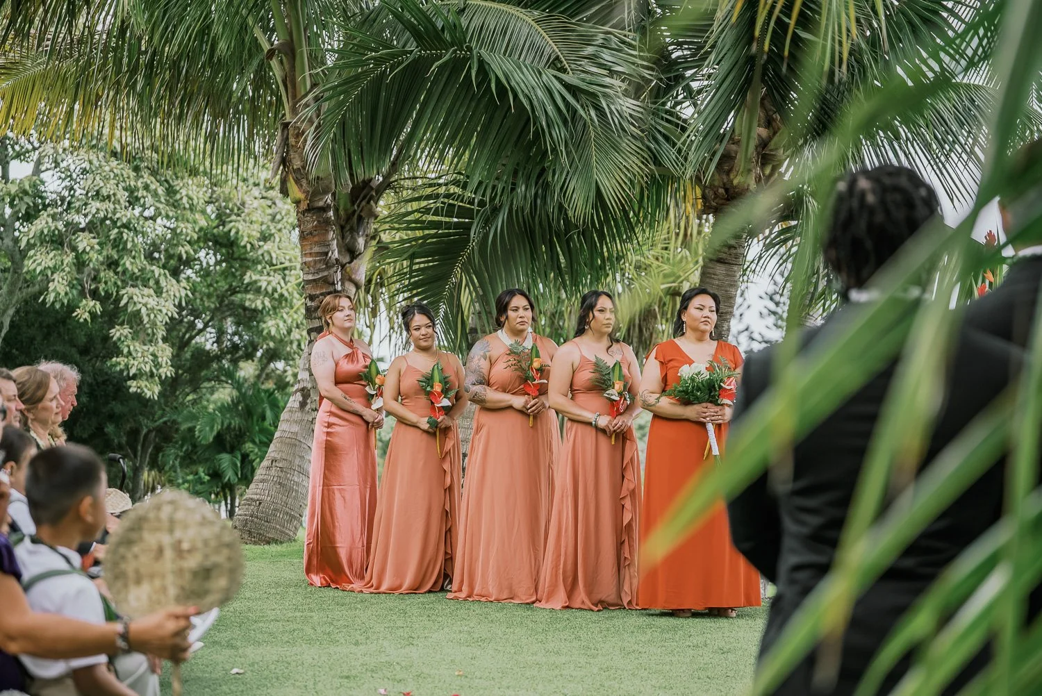 A group of five women in matching coral dresses holding floral bouquets participating in an outdoor wedding ceremony under palm trees.