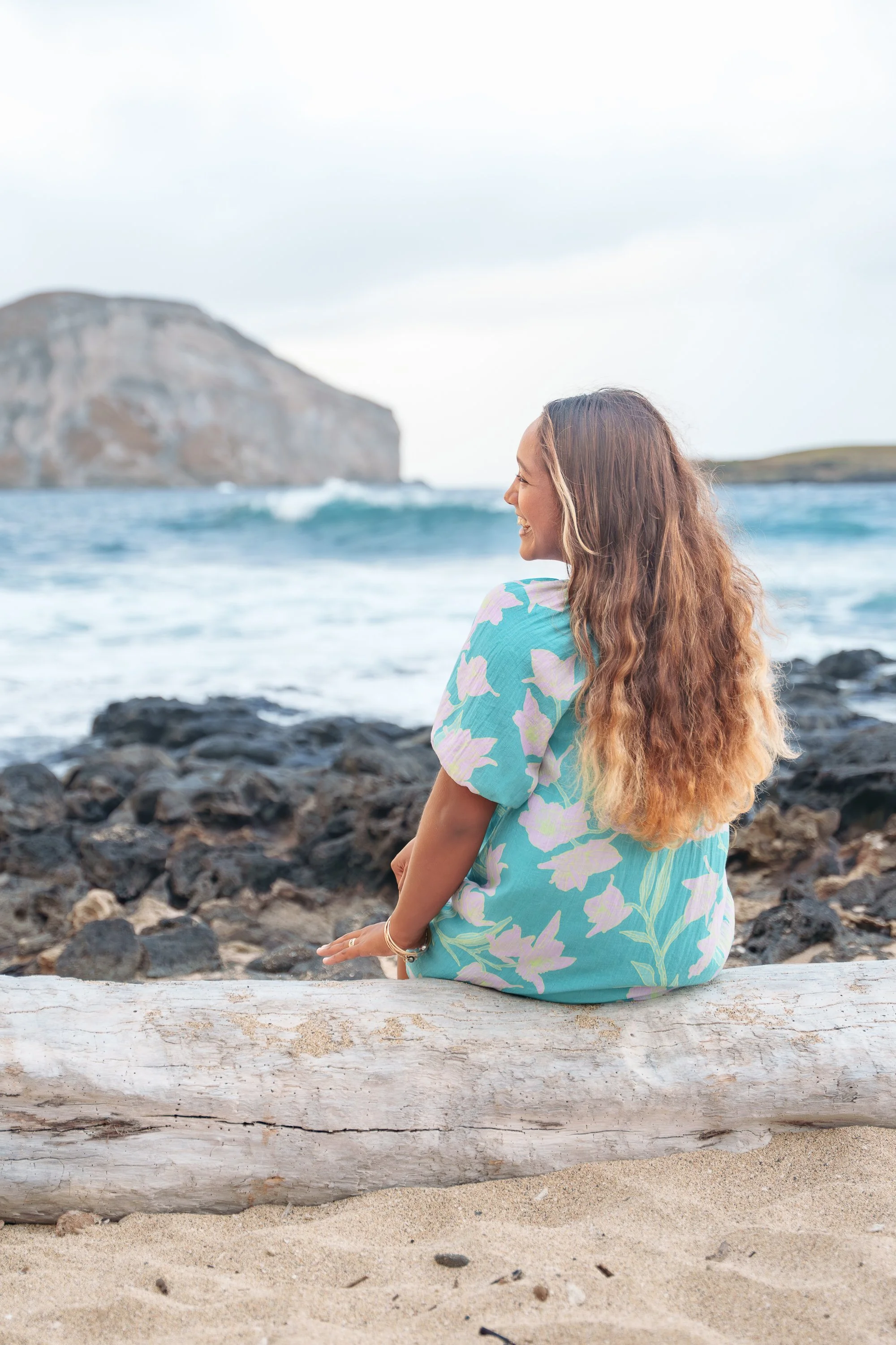 A woman with long, wavy hair sitting on a log on the sandy beach, facing the ocean with waves and cliffs in the background, smiling as she looks toward the water.