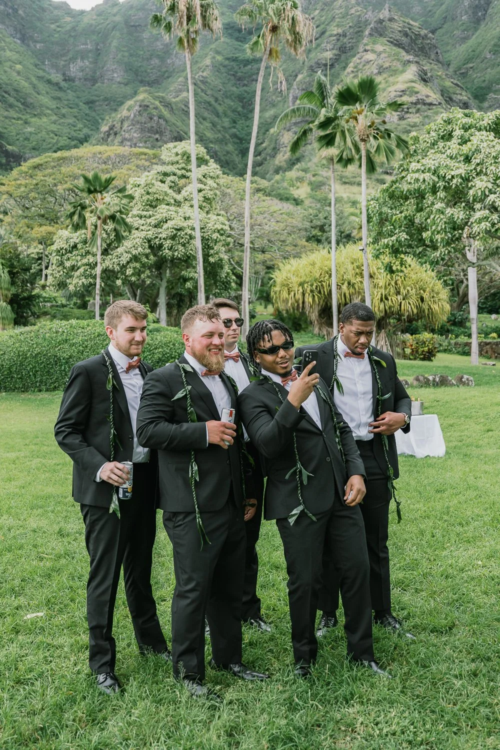 A group of five men dressed in black suits, white shirts, and bow ties stands on green grass in a lush outdoor setting with trees, palm trees, and mountains in the background. They are wearing leis made of green leaves, and some are holding drinks wh